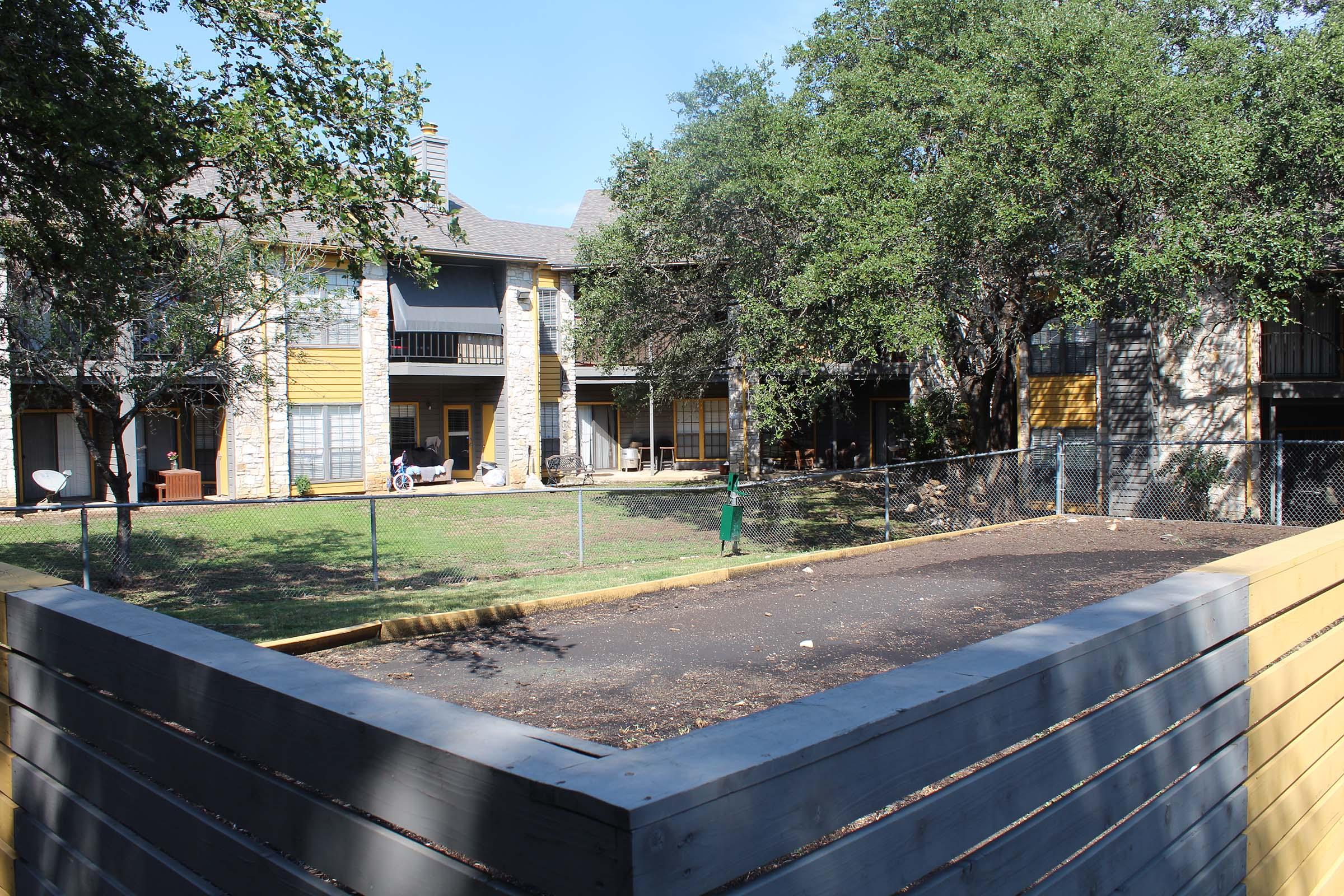A fenced outdoor area surrounded by buildings, featuring gravel ground and some trees. The background shows apartment buildings with yellow and stone exteriors, and a small grassy area. Some outdoor furniture is visible on a porch, indicating a residential setting.