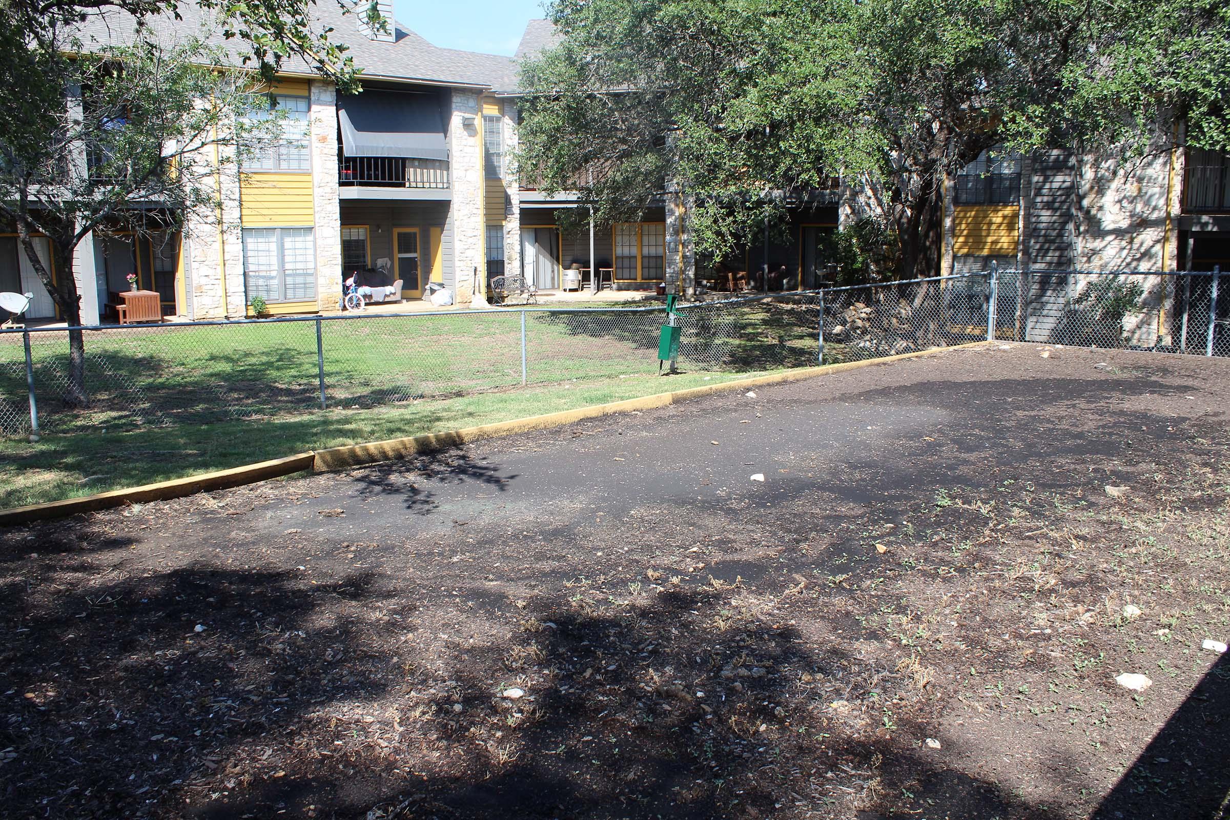 A fenced outdoor area with bare ground and some scattered debris, surrounded by trees and greenery. In the background, there are apartment buildings with balconies and patios. The space appears to be an empty yard, likely for recreational purposes.