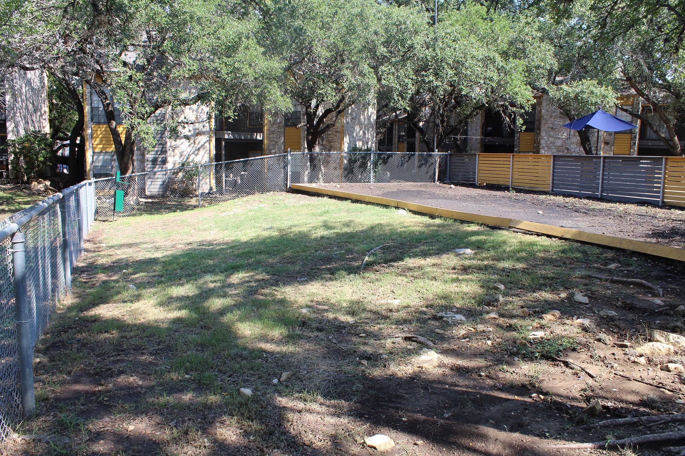 A fenced outdoor area with a wooden platform leading into a grassy space. Surrounding the area are trees and buildings, with some visible outdoor furniture, including a blue umbrella. The ground is mostly dry and uneven, indicating recent landscaping or construction work.
