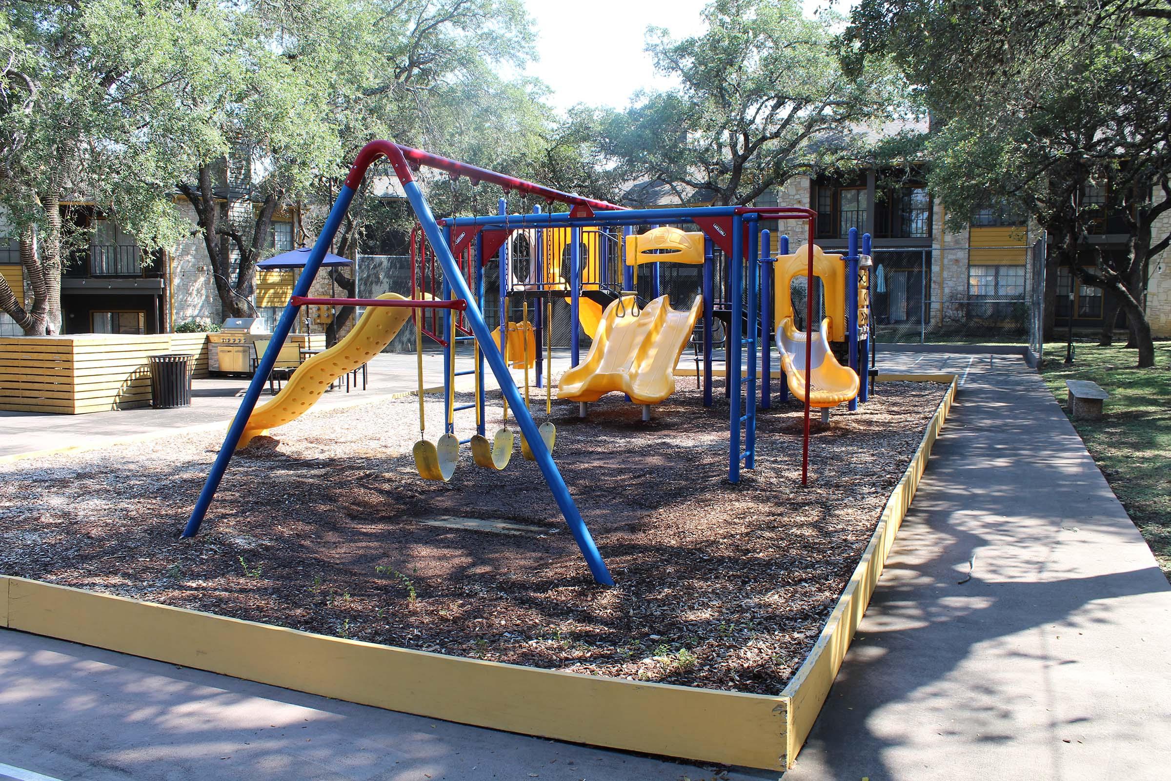 A colorful playground with blue and yellow structures, including slides and swings, situated on a gravel surface. Surrounding the playground are grassy areas and trees, with nearby buildings visible in the background. The setting is bright and inviting, designed for children's play.