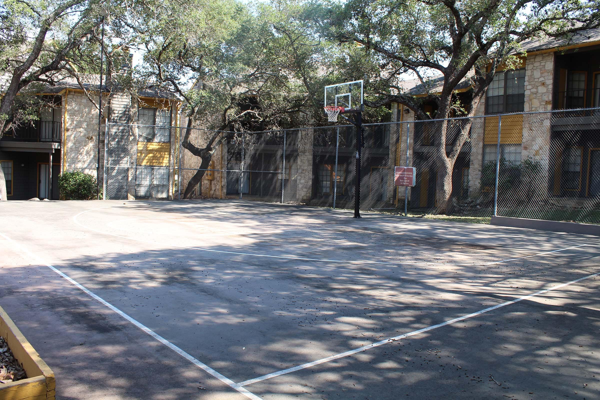 A vacant outdoor basketball court surrounded by trees and residential buildings. The court features a basketball hoop, a chain-link fence, and is mostly shaded. The surface is partially worn and has some leaves scattered on it.