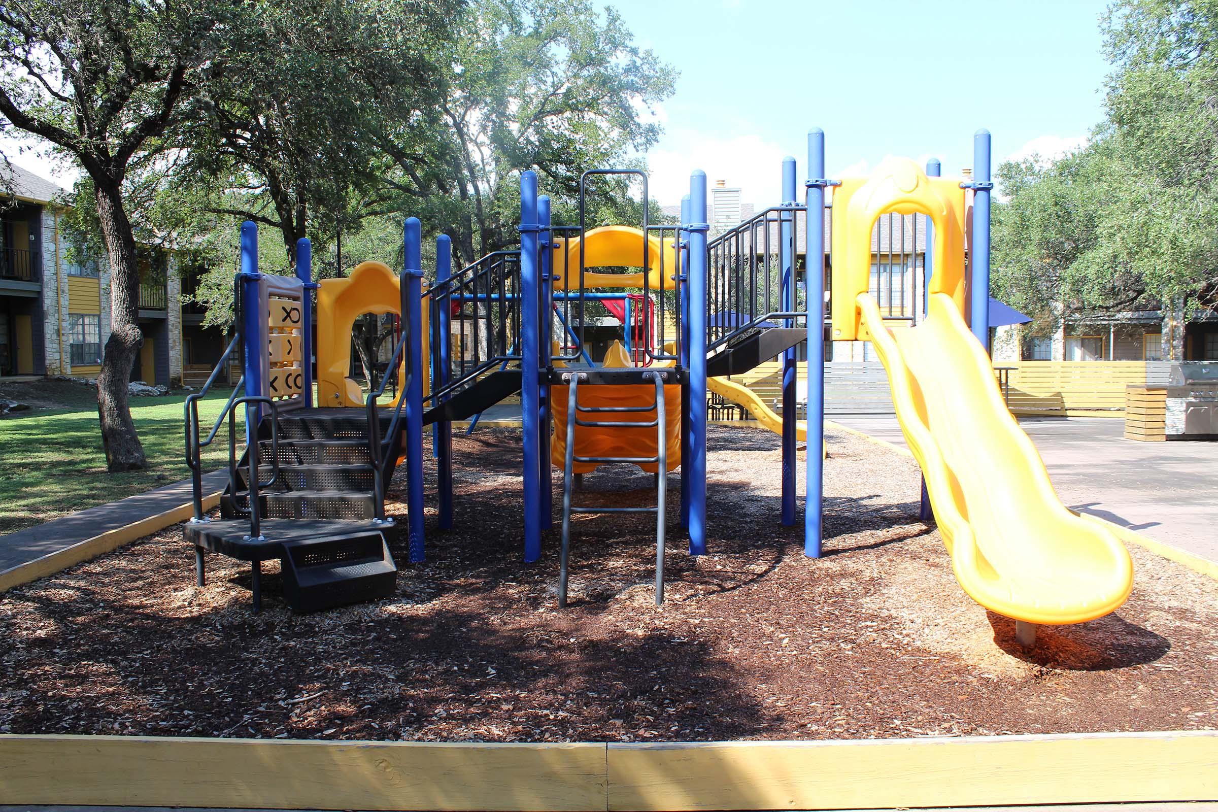 A colorful playground featuring a climbing structure, multiple slides, and swings, surrounded by trees. The play area is filled with wood chips for safety, and there are buildings in the background. The overall setting is sunny and inviting, perfect for children to play.