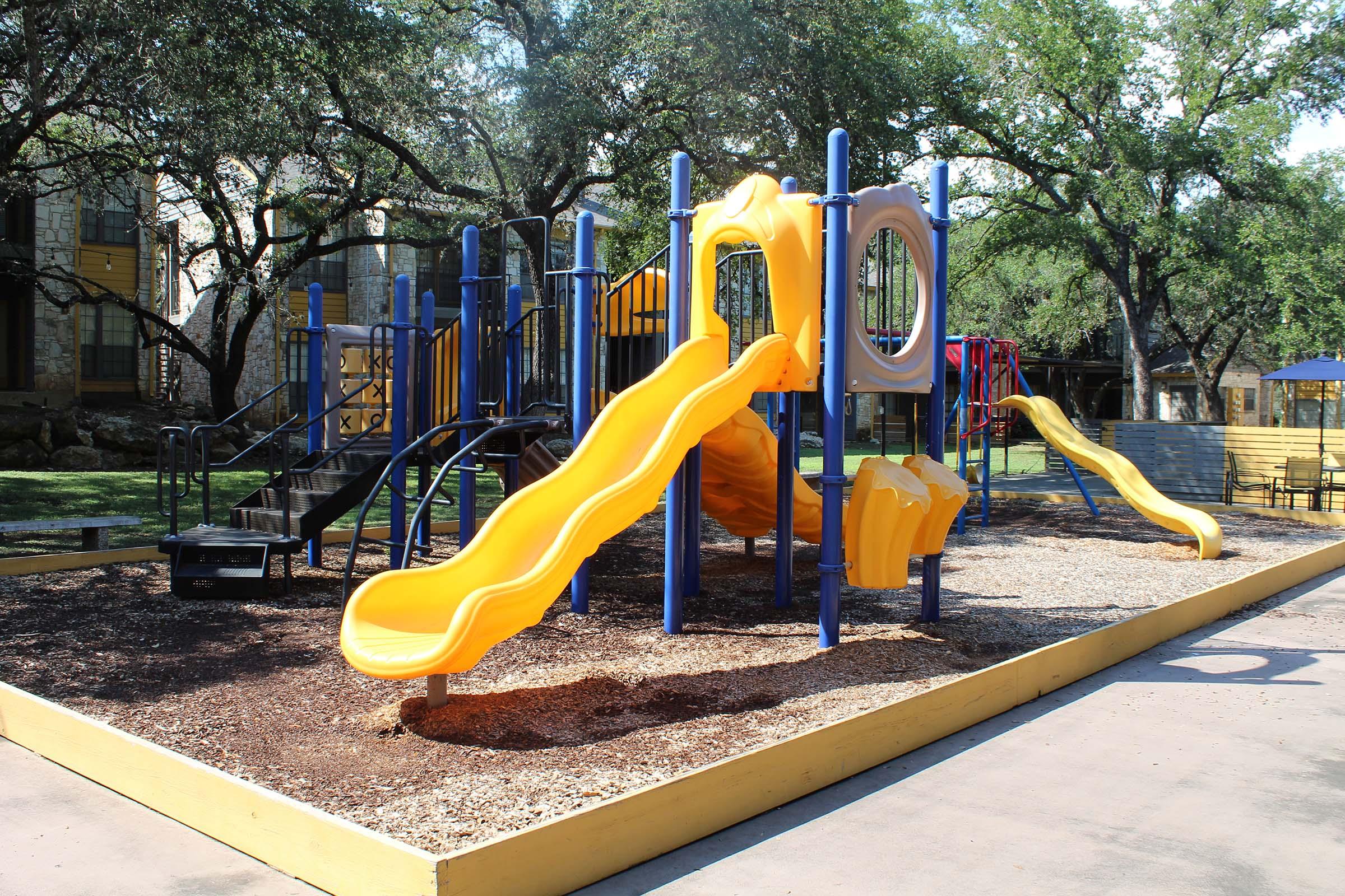Colorful playground with yellow slides, climbing structures, and shaded grassy areas. The equipment is arranged on a bed of mulch, surrounded by trees and nearby buildings, providing a fun and safe environment for children to play.
