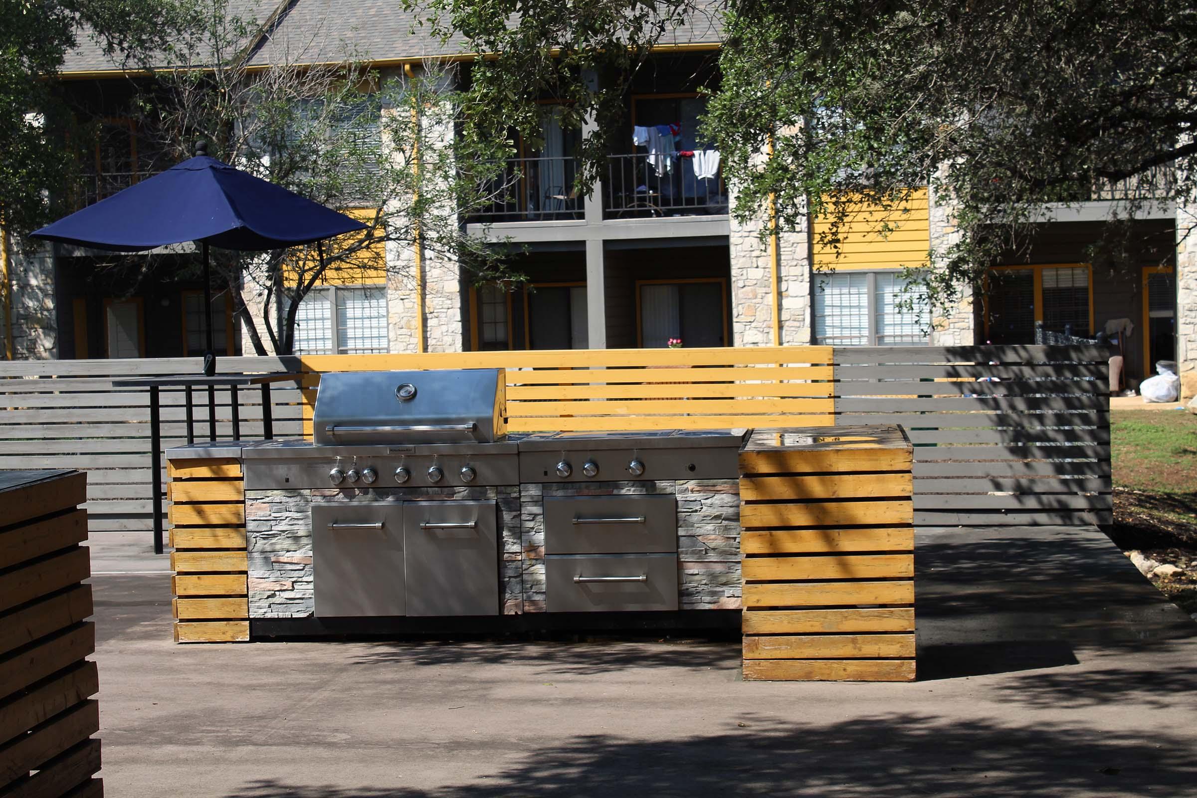 An outdoor barbecue grill with stainless steel appliances and a stone facade, situated on a patio area surrounded by wooden seating and an umbrella. In the background, there are several multi-story residential buildings with clothes drying on a balcony. Trees provide shade around the area.