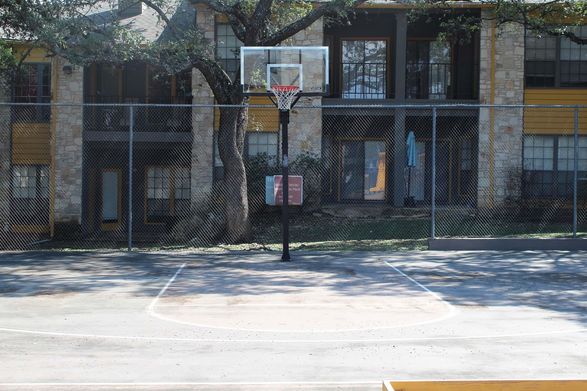 A basketball court featuring a hoop and net. The court is surrounded by a chain-link fence and has a concrete surface. In the background, there are residential buildings with large windows and greenery. The scene is bright and sunny.