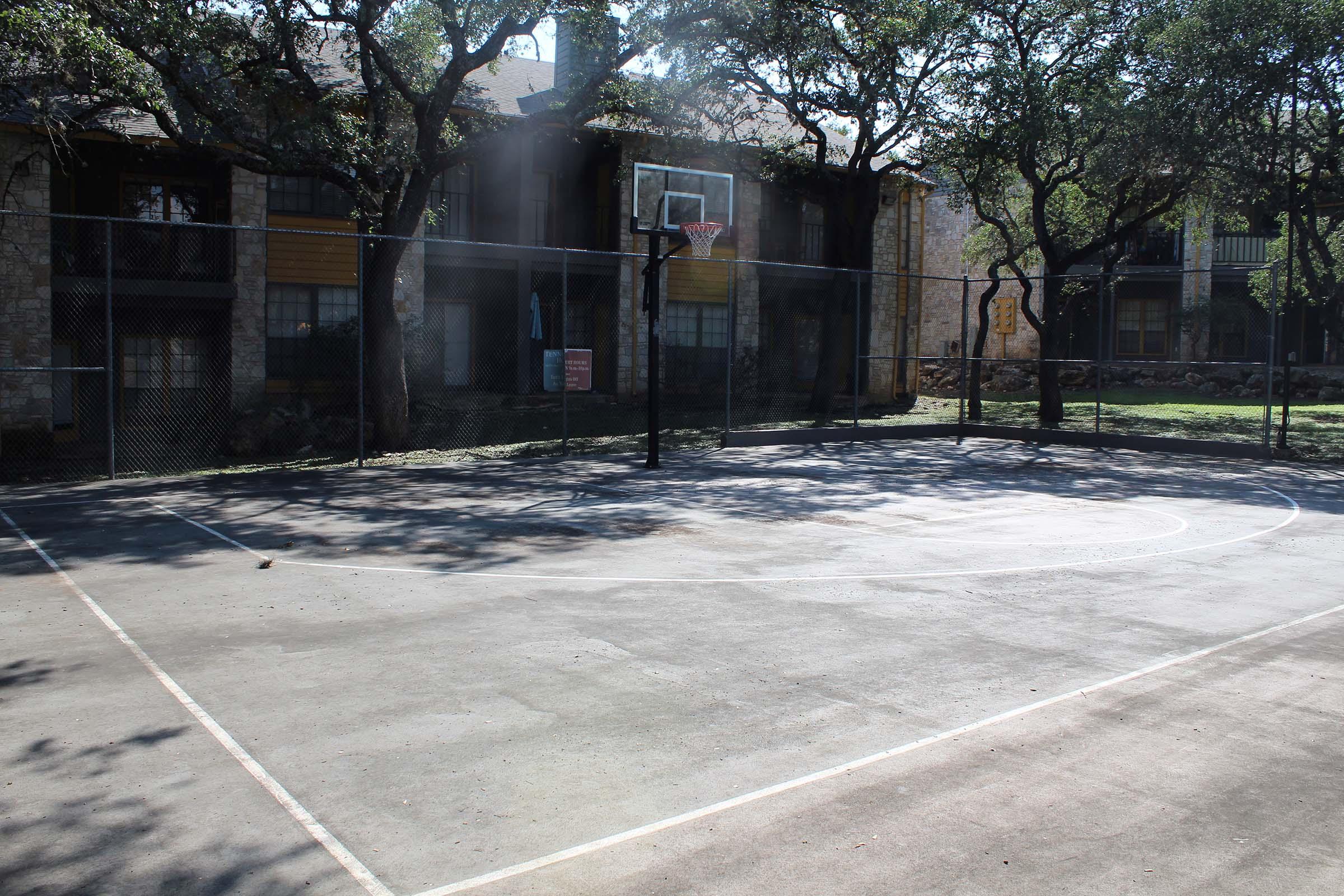 An empty outdoor basketball court surrounded by trees, with a basketball hoop at one end. In the background, there are residential buildings partially visible, framed by the foliage. Sunlight casts shadows on the court's concrete surface, highlighting the lined playing area.