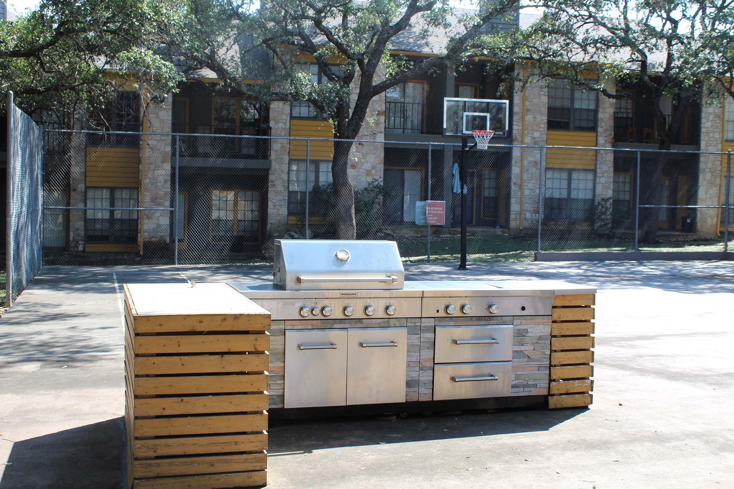 An outdoor cooking grill station made of stainless steel with wooden accents, set in a communal area near a basketball court. Behind the grill, there's a fenced playground with trees and a multi-story apartment building in the background.
