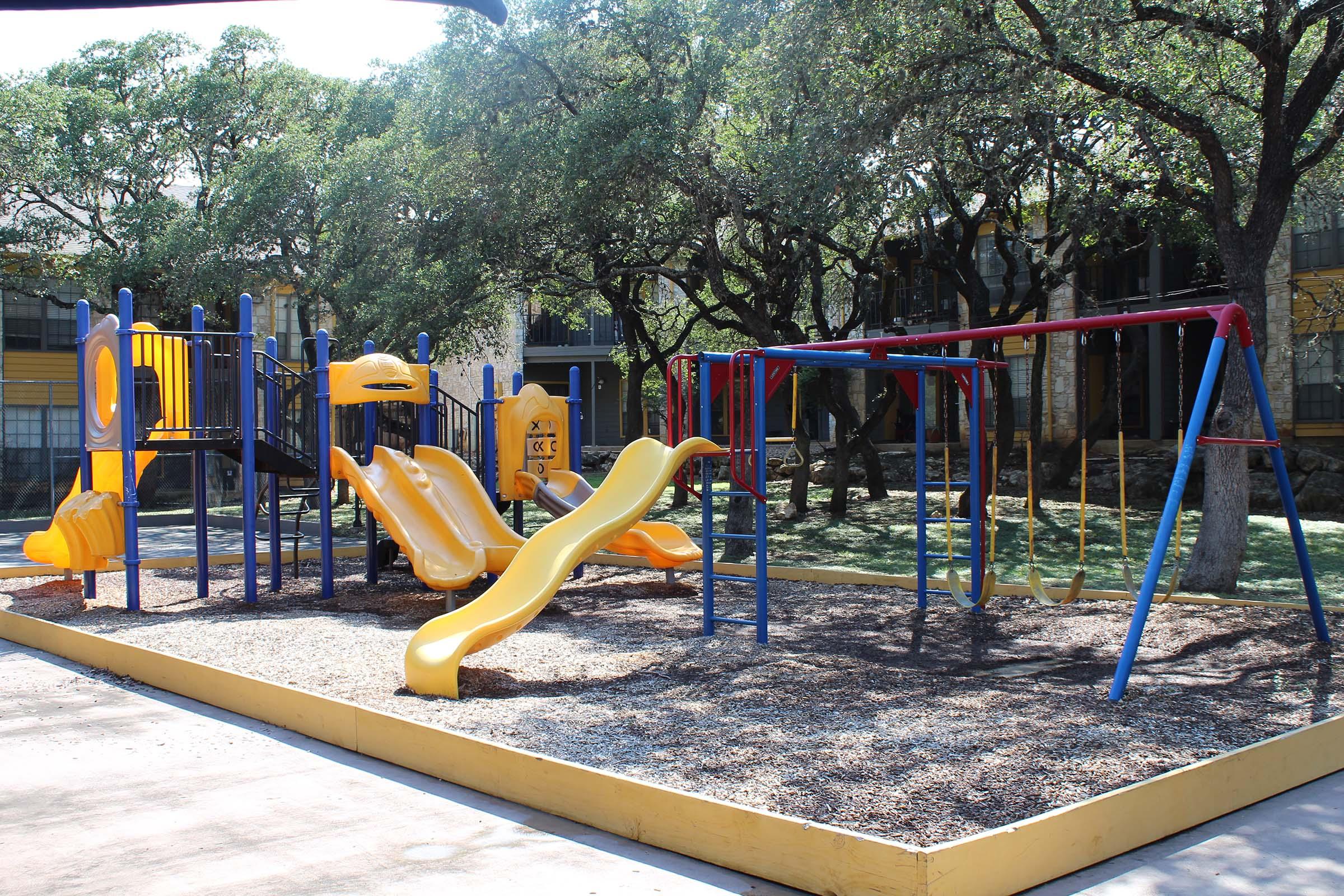 A colorful playground featuring two yellow slides, blue climbing structures, and a set of swings. The area is surrounded by trees and has a mulch surface. In the background, apartment buildings can be seen.