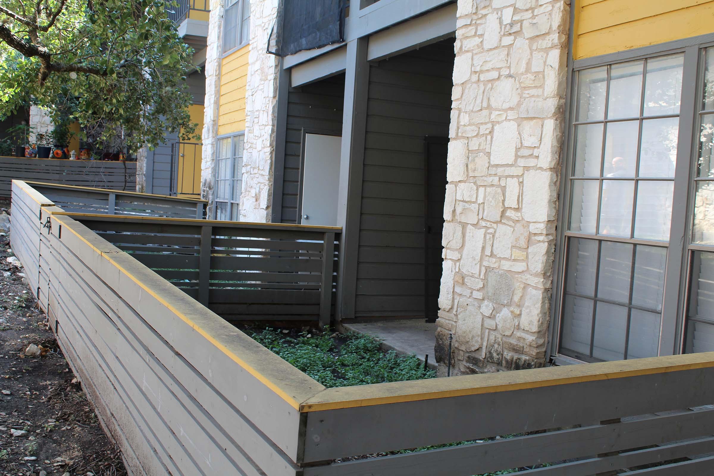 A view of a multi-story building with yellow and gray exterior walls. The image shows a partially enclosed outdoor area with wooden fencing, stone accents on the building, and green vegetation visible. There are entrances to the building on the left side, and windows with horizontal bars can be seen.