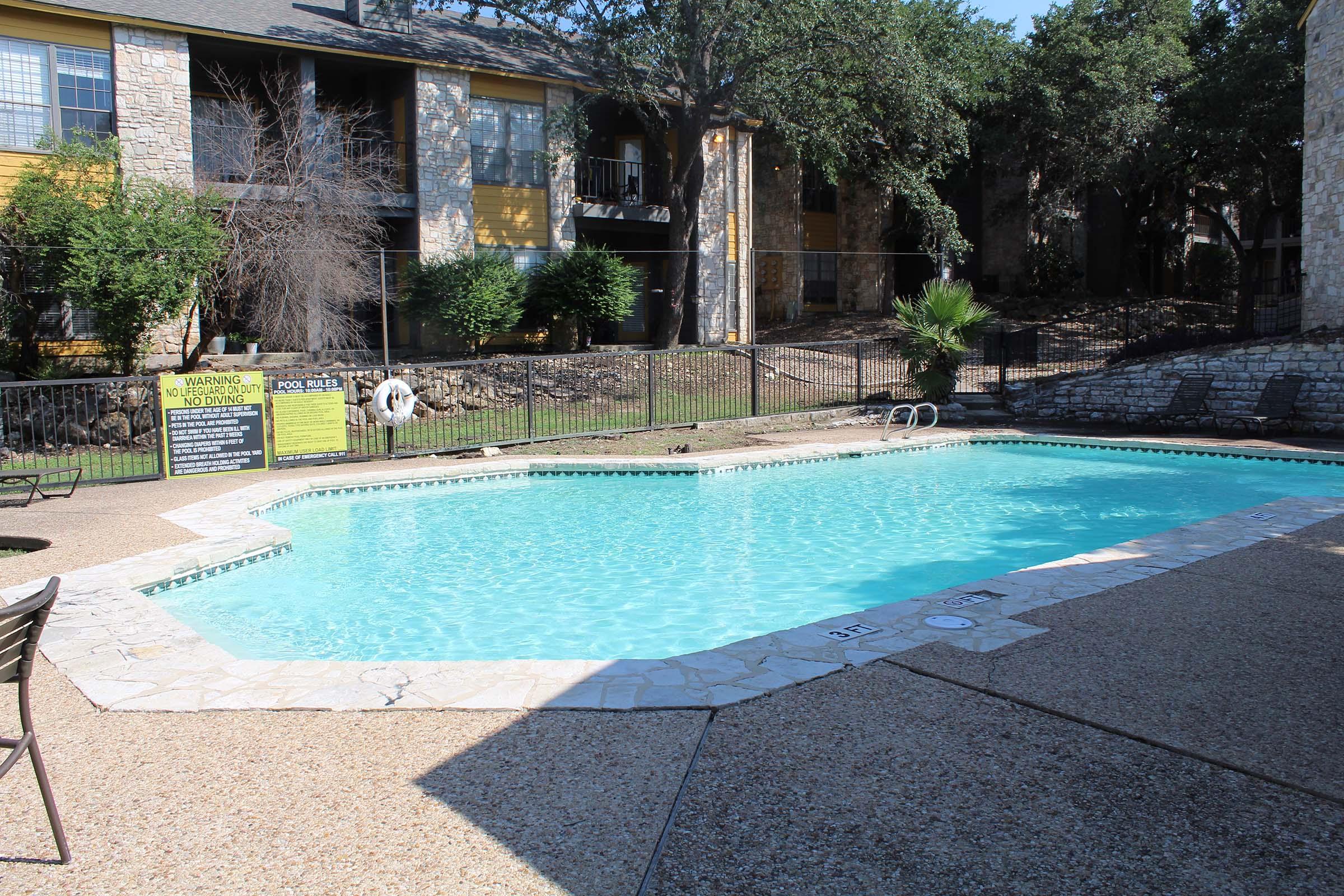 A clear blue swimming pool surrounded by a stone patio, with a warning sign posted nearby. The area is enclosed by a fence, and there are a few chairs placed around the pool. In the background, there are residential buildings partially shaded by trees.