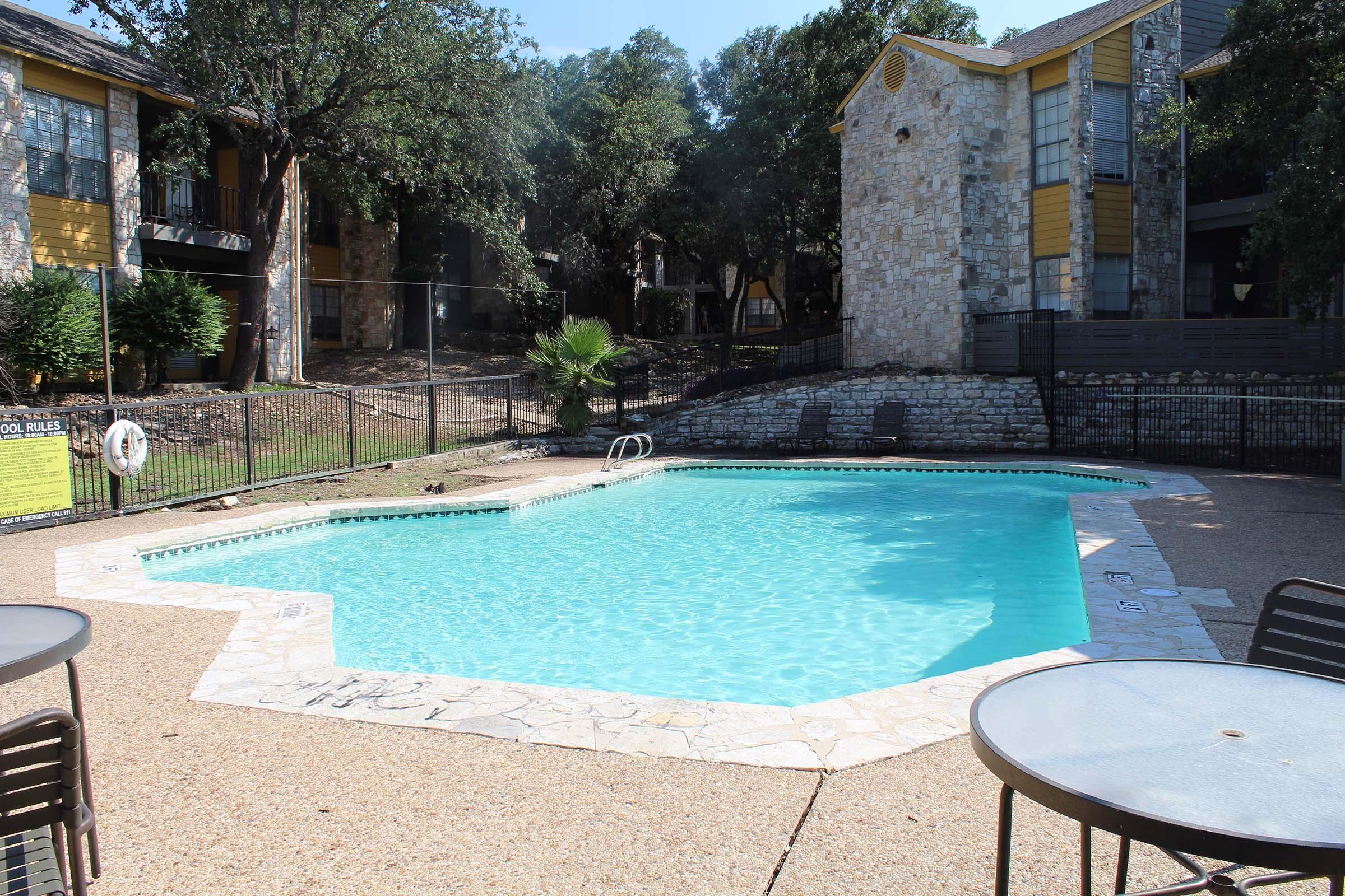 A clear blue swimming pool surrounded by a stone deck, with a ladder leading into the water. In the background, several apartment buildings can be seen, partially obscured by trees. There are patio tables with chairs nearby, and a fence enclosing the pool area.
