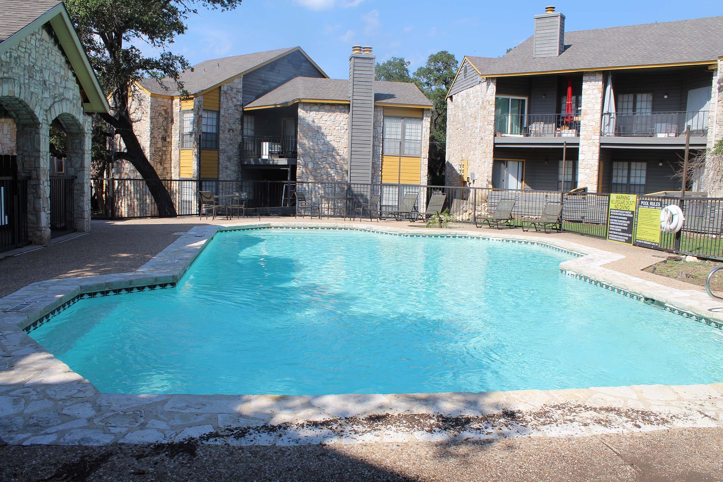 A clean, inviting swimming pool surrounded by a stone deck, located in an apartment complex. There are two buildings visible in the background, featuring balconies and a mix of stone and siding exteriors. Lush green trees provide shade nearby under a clear blue sky.