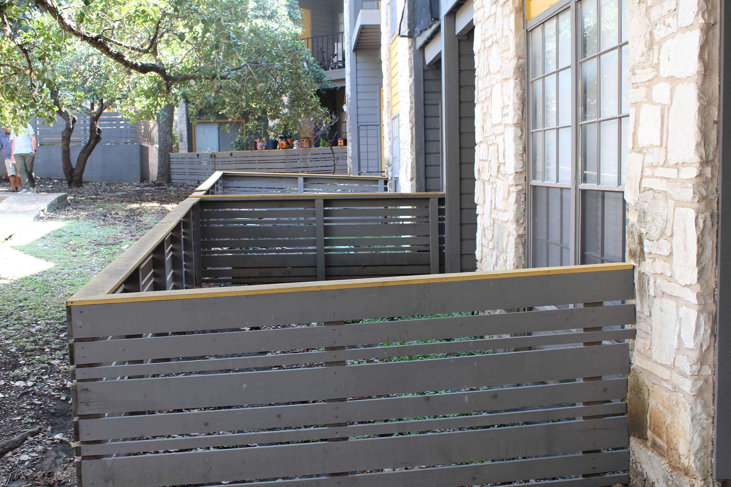 A view of a gray wooden railing surrounding a small outdoor area adjacent to a building. The setting is surrounded by trees. In the background, some blurred figures of people can be seen near the building, adding a sense of liveliness to the scene.