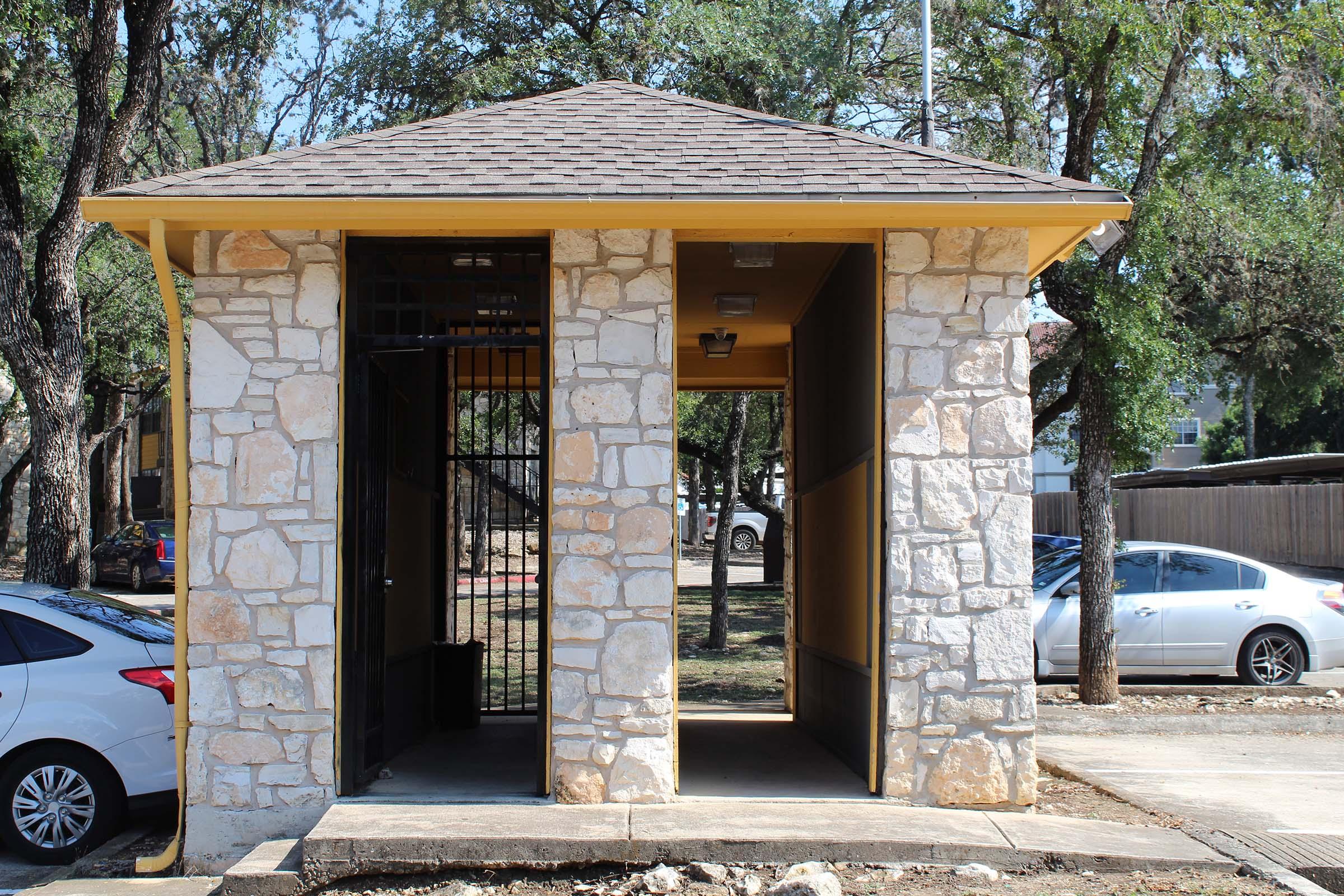 A small, open-sided building with a stone exterior and a yellow roof, situated in a park-like area. One side features a gated entrance, while the other is open. Surrounding the building are trees and parked cars, giving a glimpse of a residential or recreational setting.