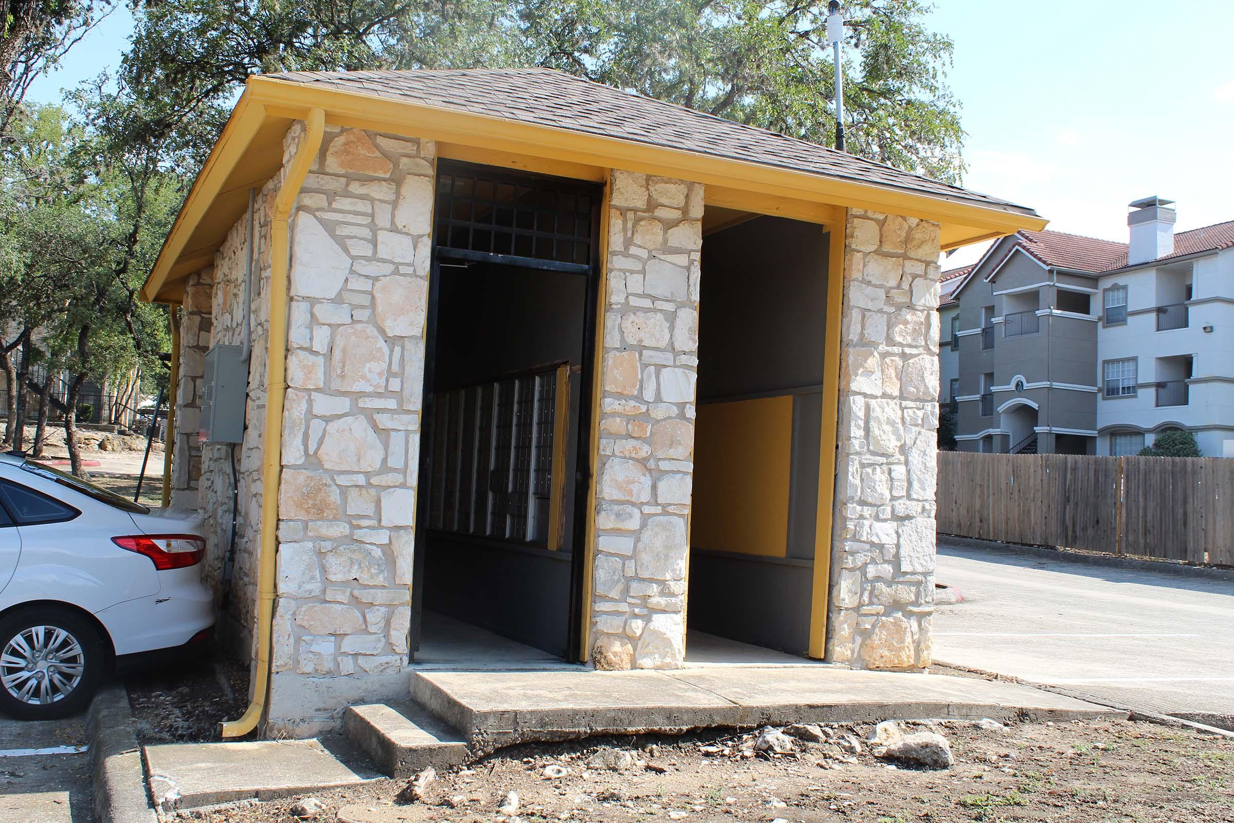 A small, stone building with a yellow roof and trim. The structure has two open entrances, with a pathway leading up to it. Nearby, a white car is parked, and in the background, there are apartment buildings behind a wooden fence. Trees are visible around the area.