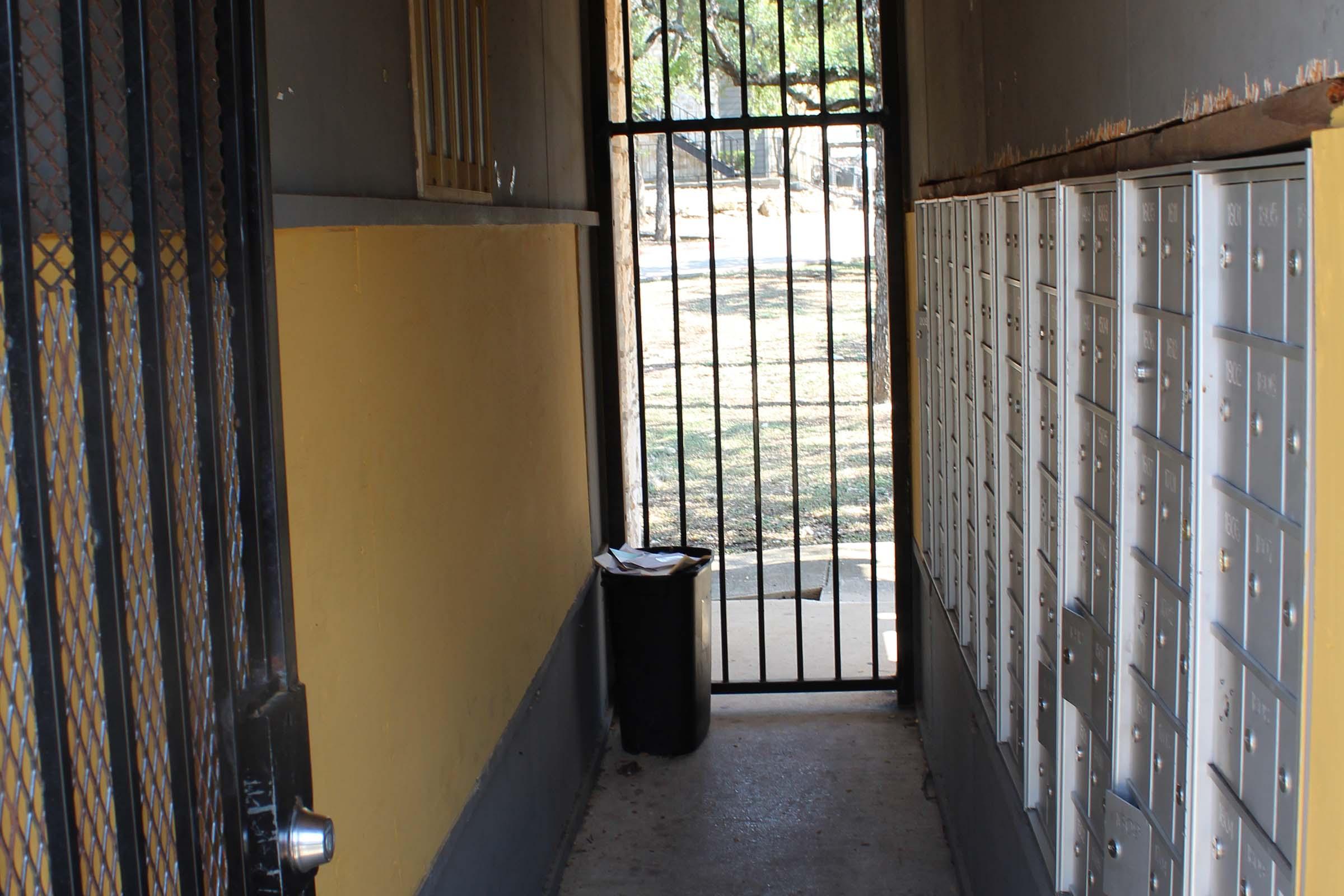 A narrow corridor leading to a gated area with mailbox units on one side. A black trash can is located at the end of the hallway, and the outside view shows a grassy area beyond the gate. The walls are painted in yellow and gray.