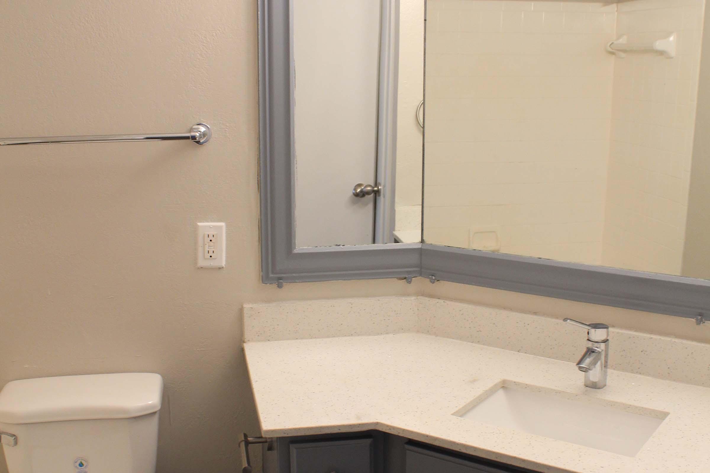 A clean, modern bathroom featuring a light-colored countertop with a sink, a mirror framed in gray, a toilet, and a hand towel rack. The background includes a shower area with white tiles, contributing to a minimalist aesthetic.