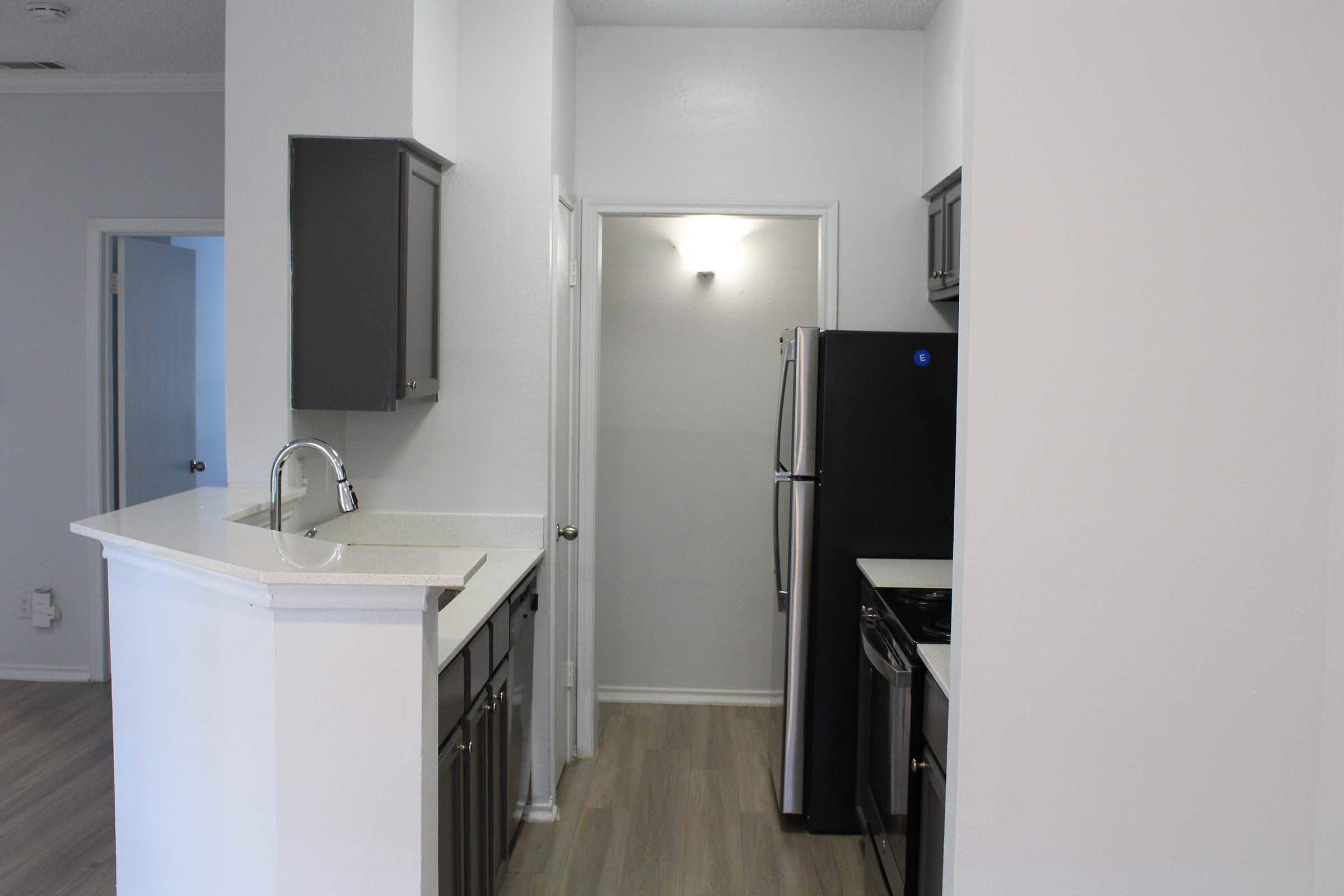 A modern kitchen view featuring a counter with a sink, gray cabinetry, a black refrigerator, and an adjoining hallway illuminated by a ceiling light. The flooring is light-colored wood laminate, and the walls are painted in a neutral shade, creating a clean and spacious atmosphere.