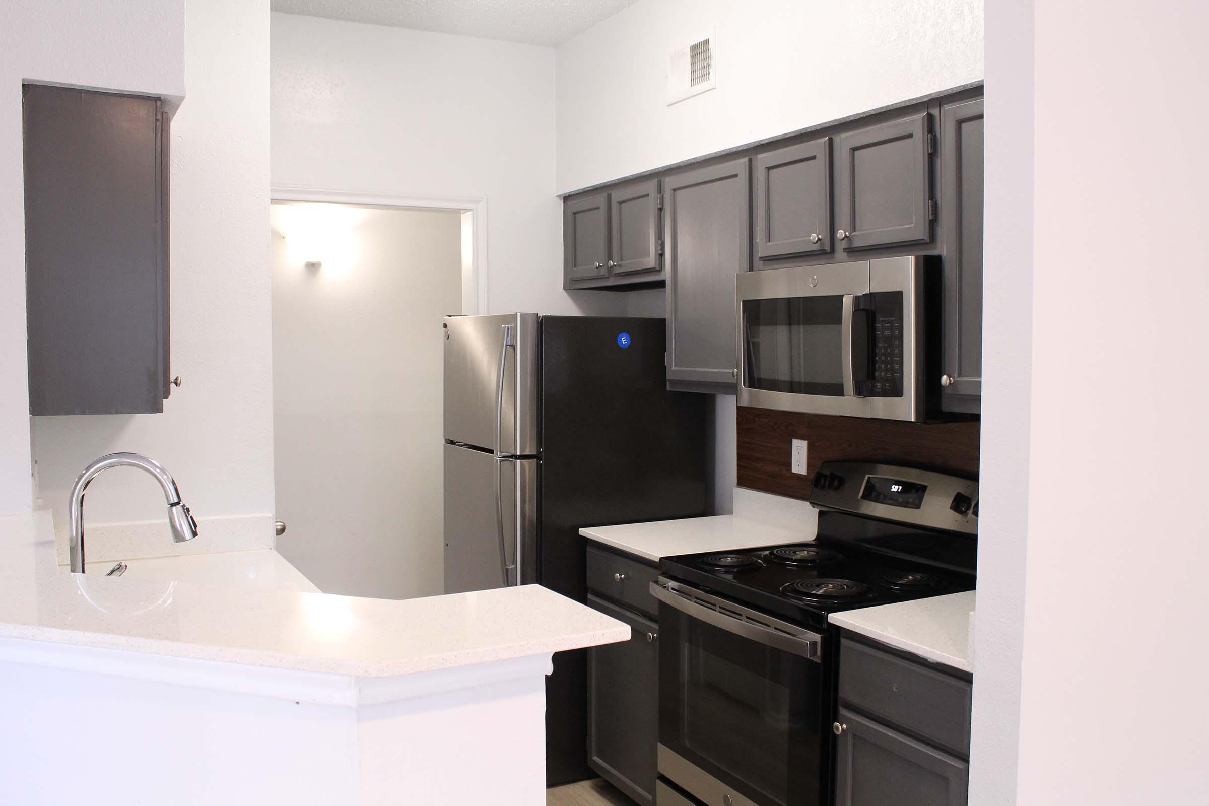 Modern kitchen featuring dark cabinetry, stainless steel appliances including a refrigerator, microwave, and oven, alongside a white countertop. The open layout includes a breakfast bar and light-colored walls, creating a spacious and inviting atmosphere.