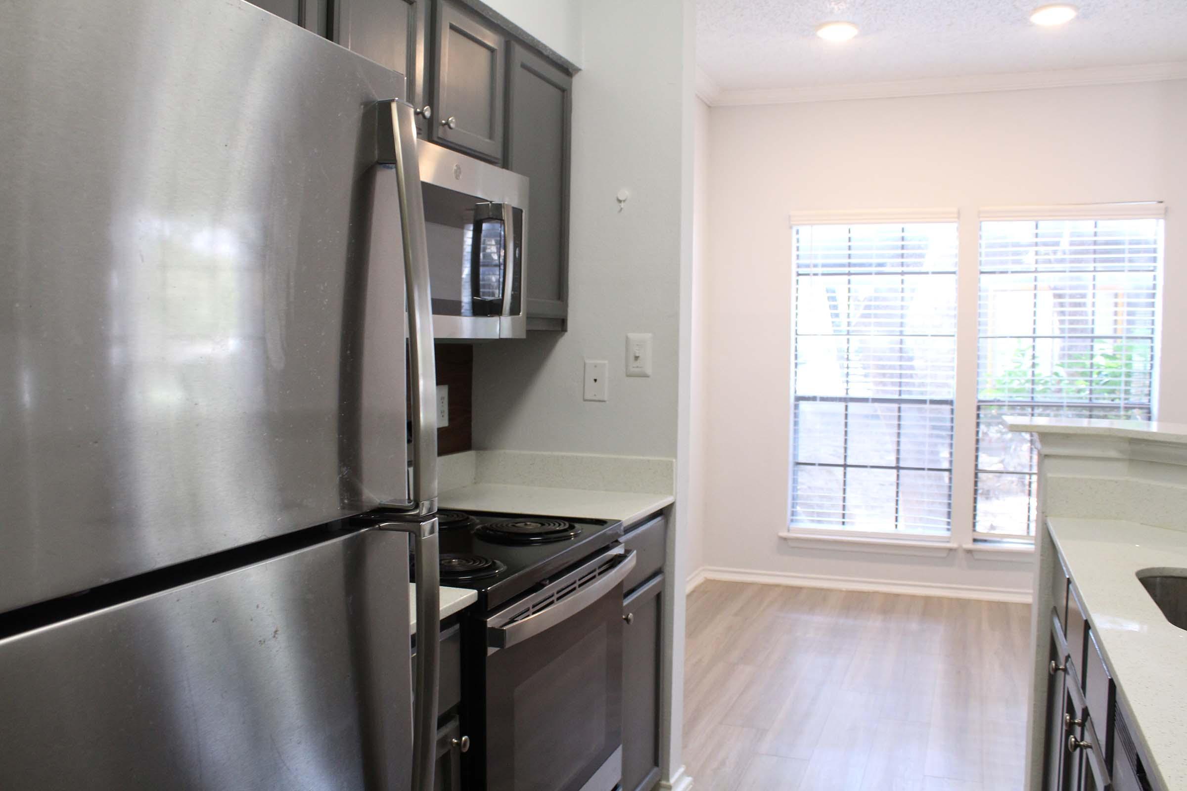 Modern kitchen interior featuring stainless steel appliances, including a refrigerator and oven. Light-colored countertops and cabinetry complement the space, with a window allowing natural light to brighten the area. The flooring is a light wood-like material, enhancing the contemporary design.