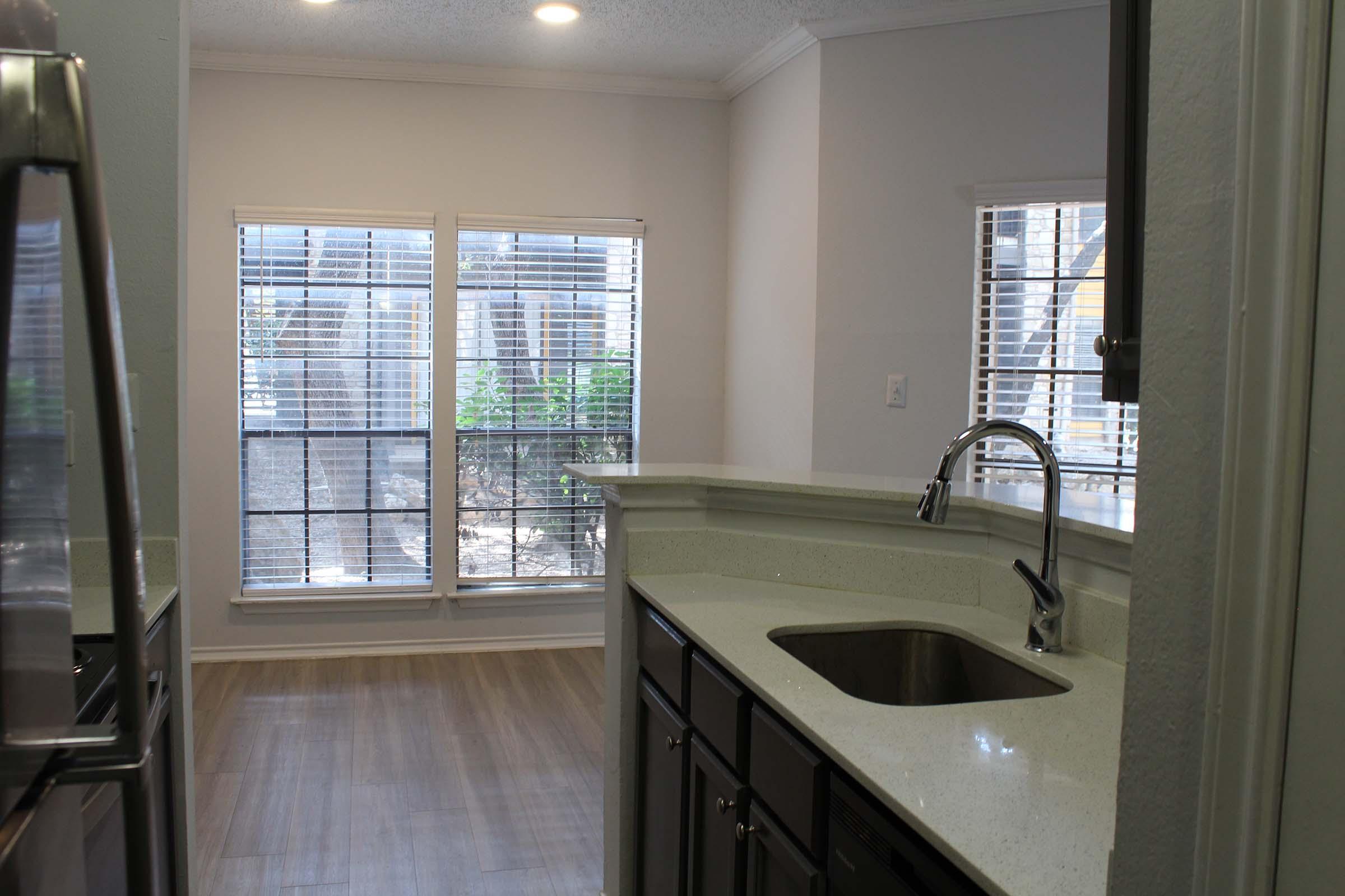 Bright and modern kitchen view with a sleek countertop and stainless steel sink, leading to a living area featuring large windows with blinds. The space has light-colored walls and wooden flooring, creating an airy atmosphere with natural light coming in from outside.