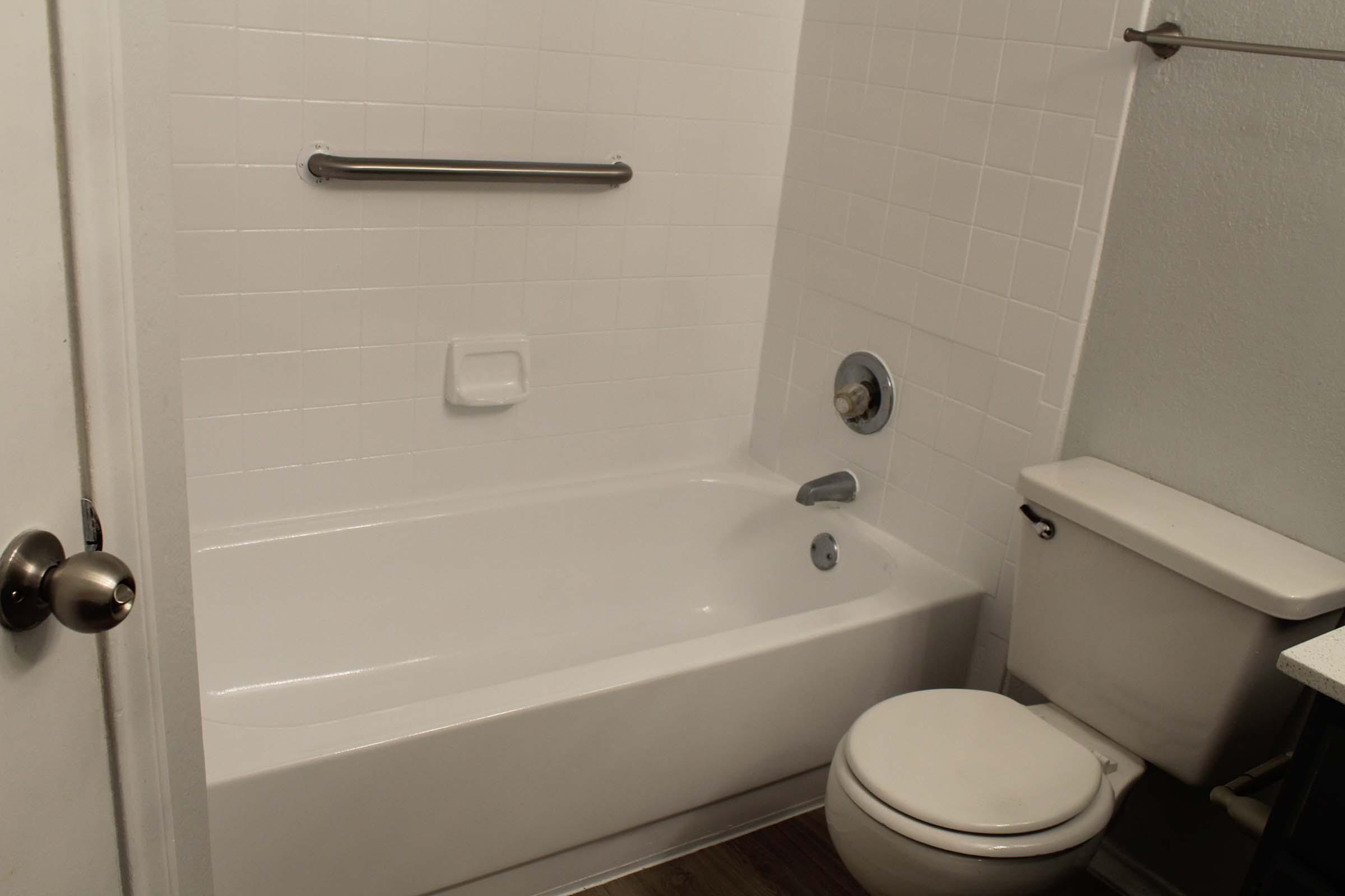 A clean, simple bathroom featuring a white bathtub with a showerhead, a chrome grab bar on the wall, and a white toilet. The walls are tiled in white, and the overall design is minimalistic with a light, modern aesthetic.