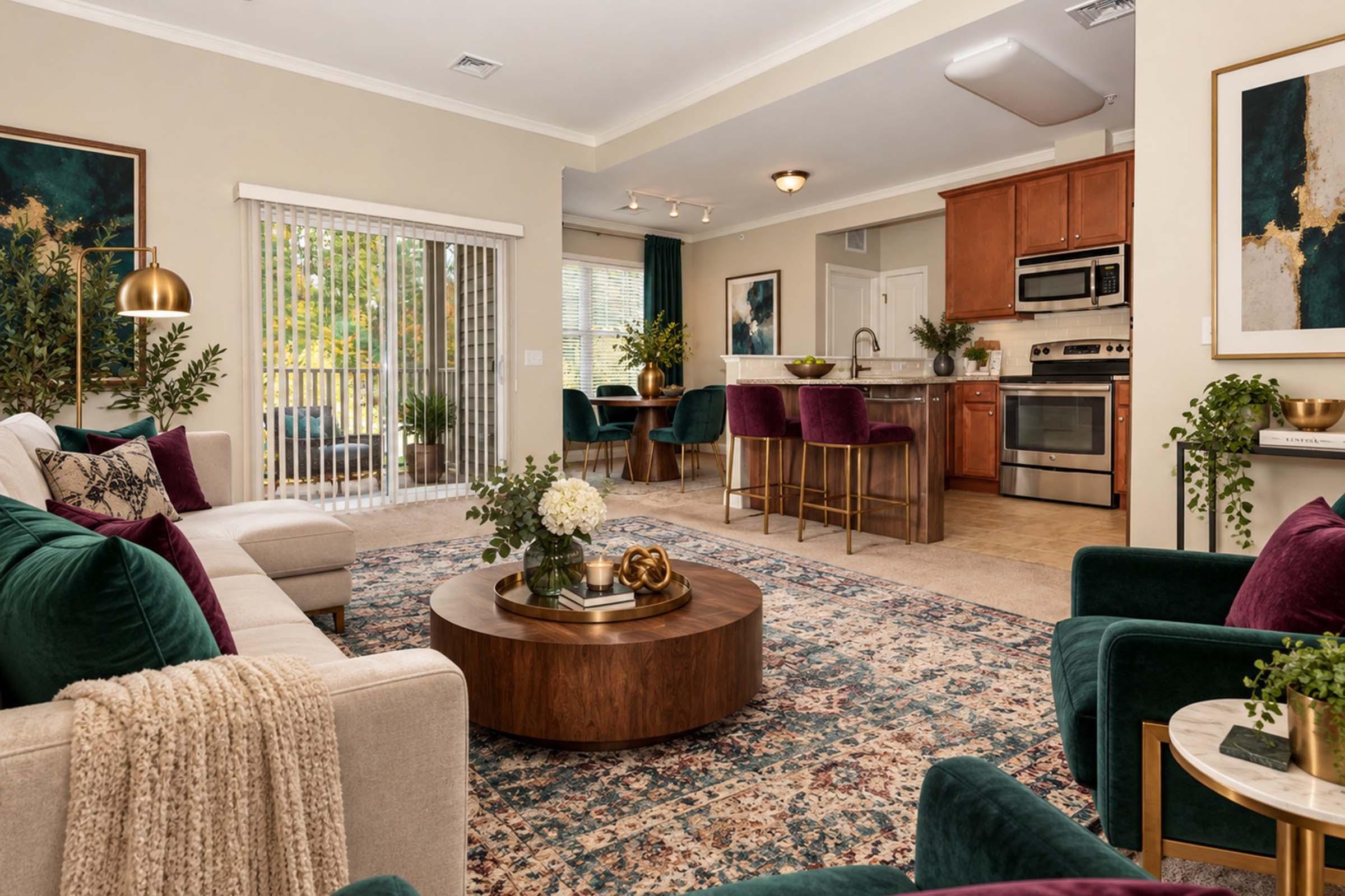A modern living room featuring a cozy sofa with plush cushions, a large area rug, and a round wooden coffee table. The space includes a dining area with bar stools, an adjacent kitchen with stainless steel appliances, and large windows allowing natural light. Decor elements include green plants and contemporary artwork.