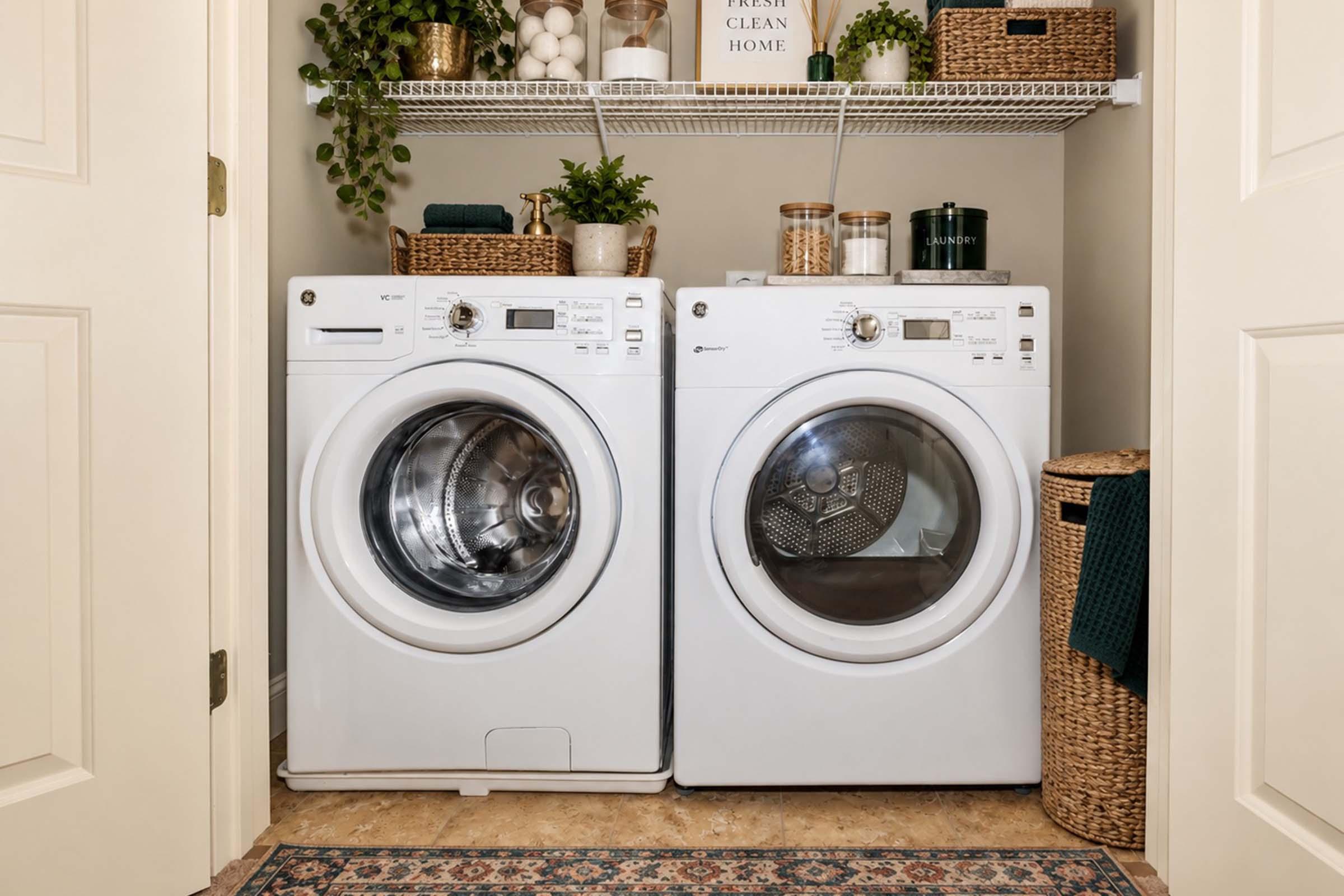 A clean laundry area featuring a stacked washer and dryer in white, surrounded by plants and storage containers. A woven basket is placed nearby, and a patterned rug adds warmth to the space. The shelves above hold decorative items and supplies, creating a tidy, organized look.