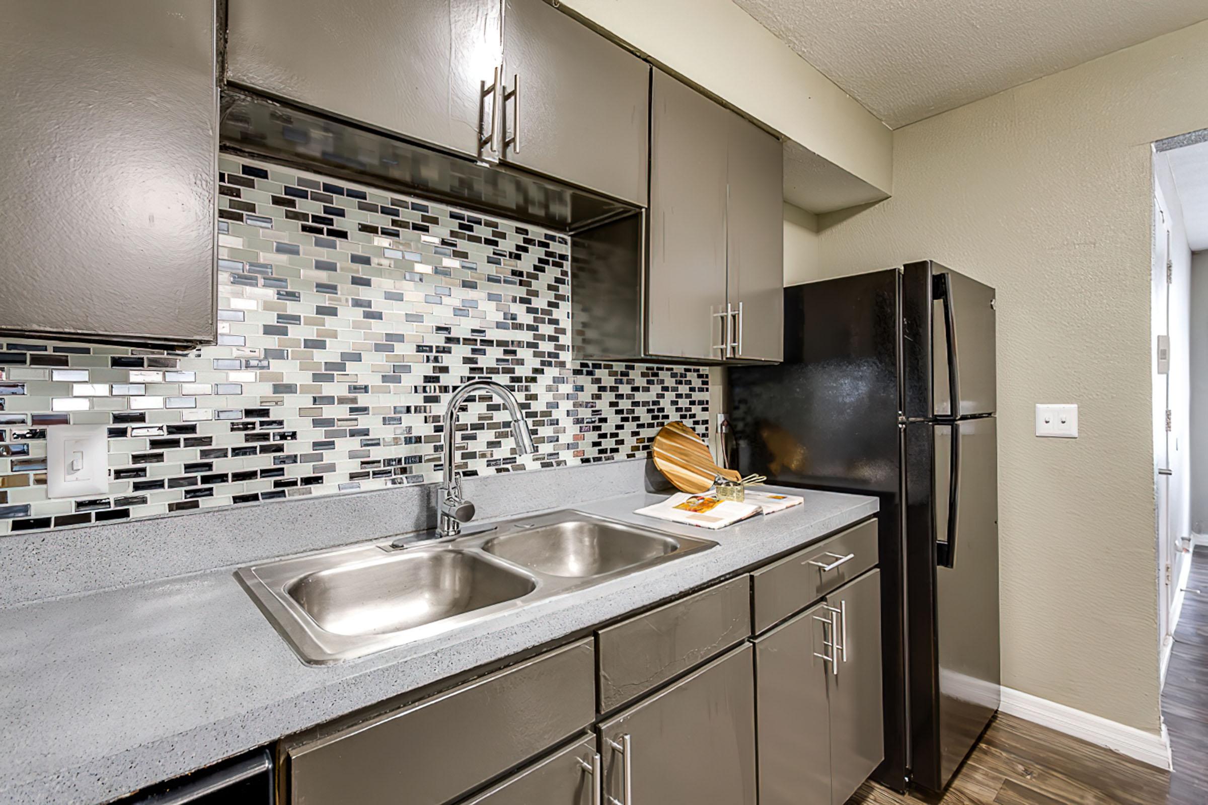 Modern kitchen featuring dark cabinetry, a stainless steel sink, and a grey countertop. A mosaic tile backsplash in black and silver tones adds visual interest. A black refrigerator is on the right, and a wooden cutting board sits next to the sink. The flooring appears laminate, complementing the cabinet colors.