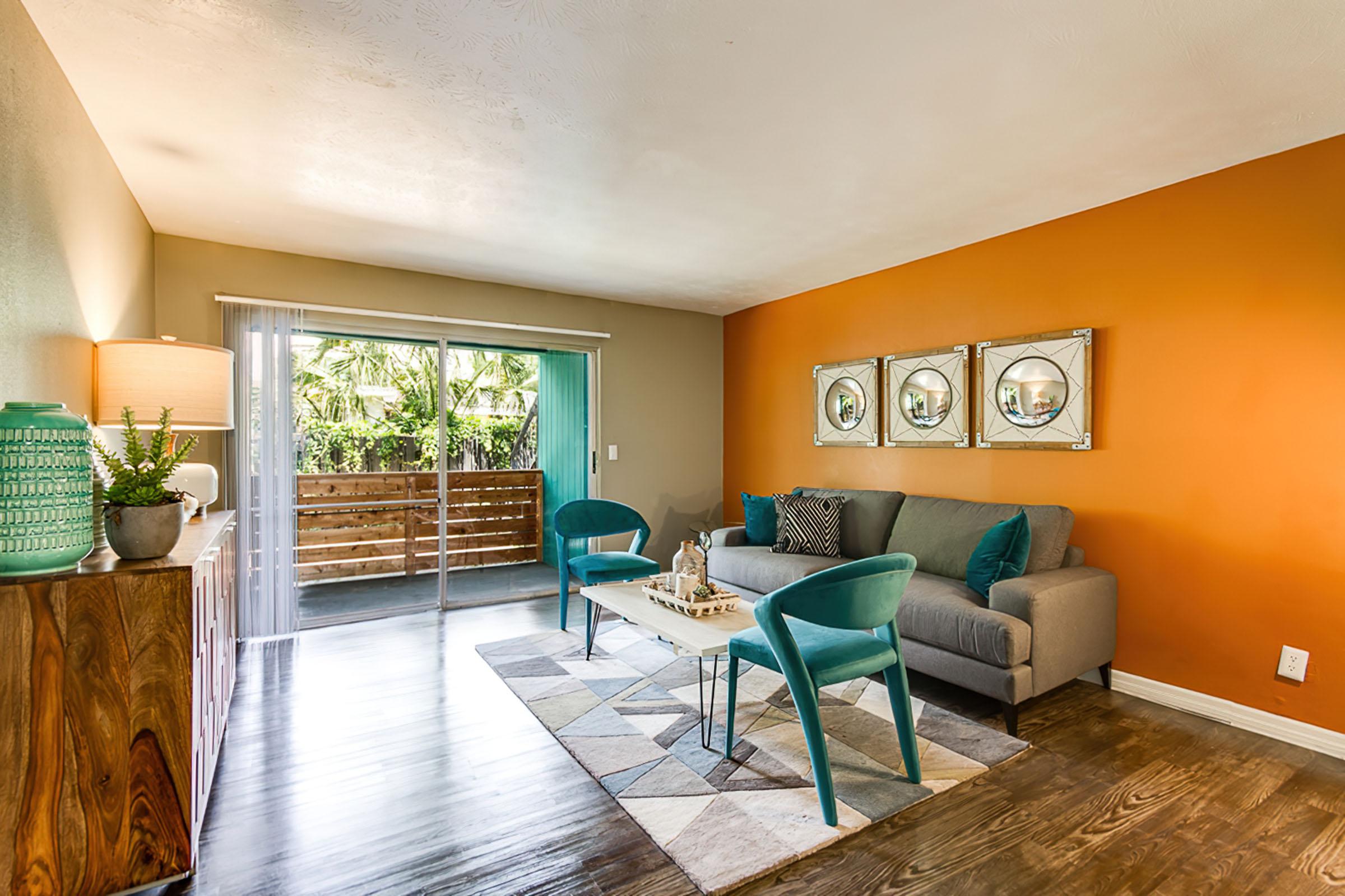 A stylish living room featuring a gray sofa with decorative pillows, teal chairs, and a light wood coffee table. The decor includes circular mirrors on an orange accent wall, a lamp on a wooden cabinet, and sliding glass doors leading to a balcony with greenery. The flooring is dark wood, enhancing the cozy atmosphere.
