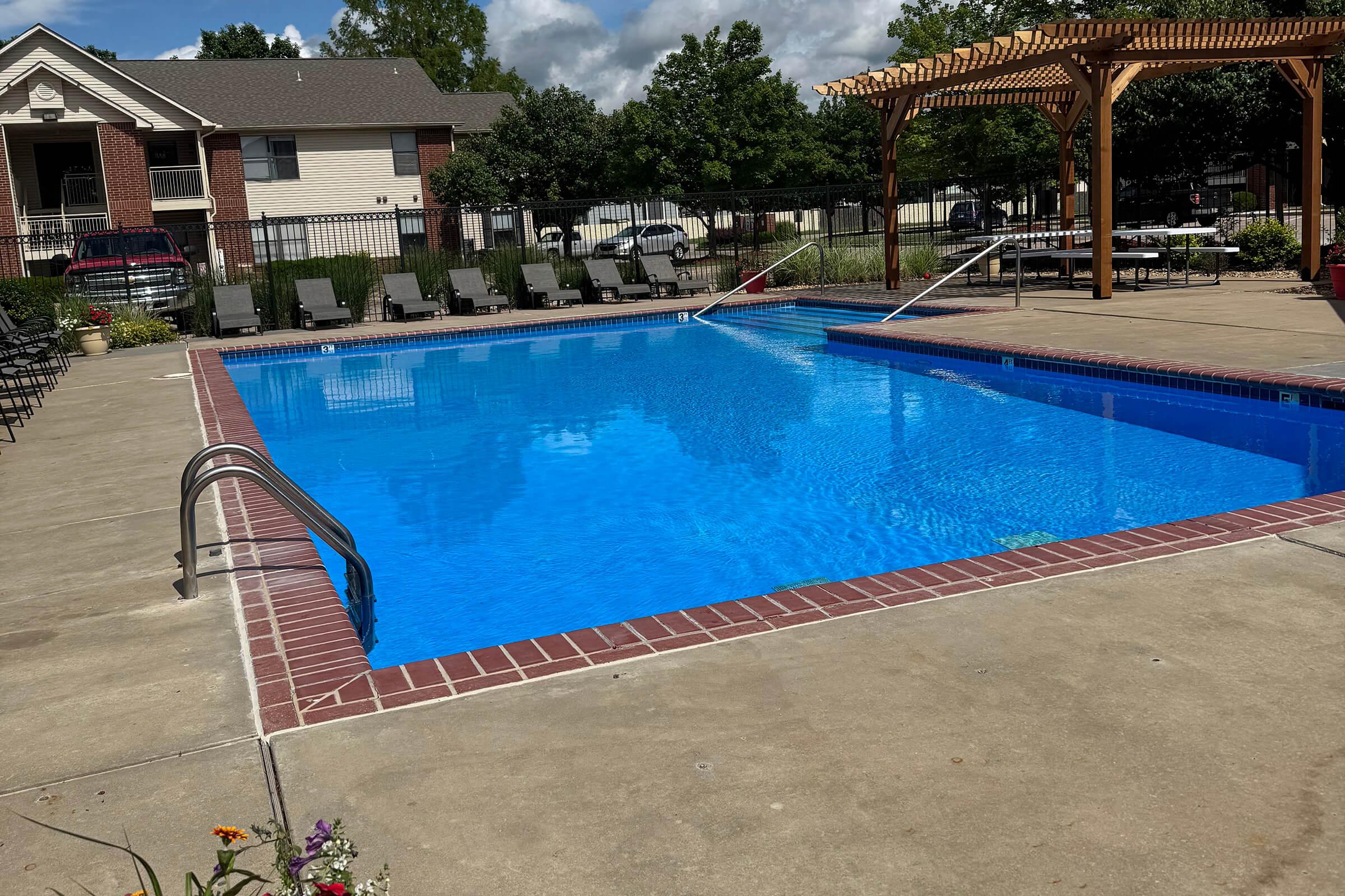 A clear blue swimming pool surrounded by a patio with lounge chairs, a pergola, and landscaped greenery. The sky is partly cloudy, and there are tables in the background, suggesting a recreational area.