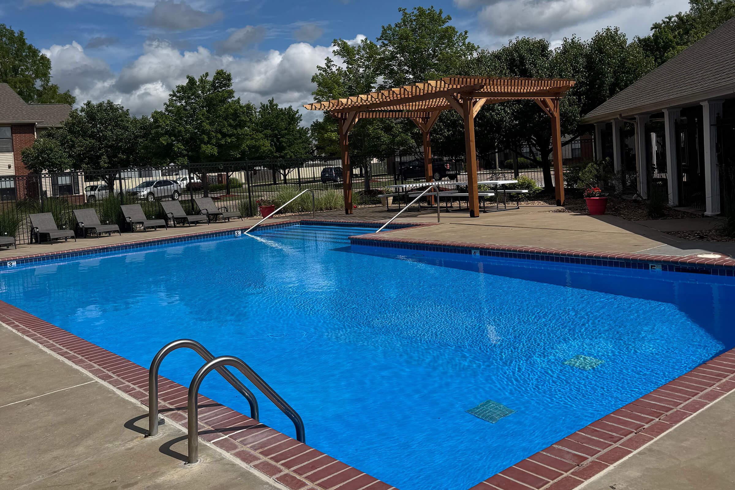 A clear blue swimming pool surrounded by a concrete deck, featuring a wooden pergola in the background. Lounge chairs are arranged along the poolside, and trees and greenery are visible in the surrounding area under a partly cloudy sky.