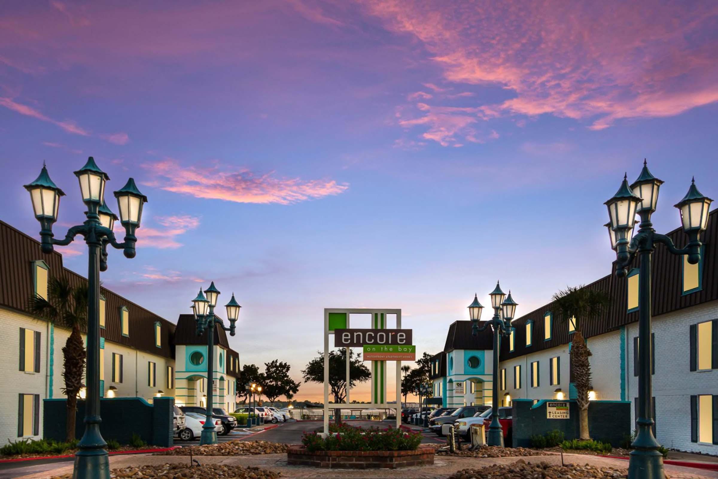 A hotel entrance at sunset featuring a large sign that reads "encore," flanked by decorative lamp posts. The building has a modern design with blue accents and palm trees in the background, providing a welcoming atmosphere. The sky is painted with hues of pink and purple.