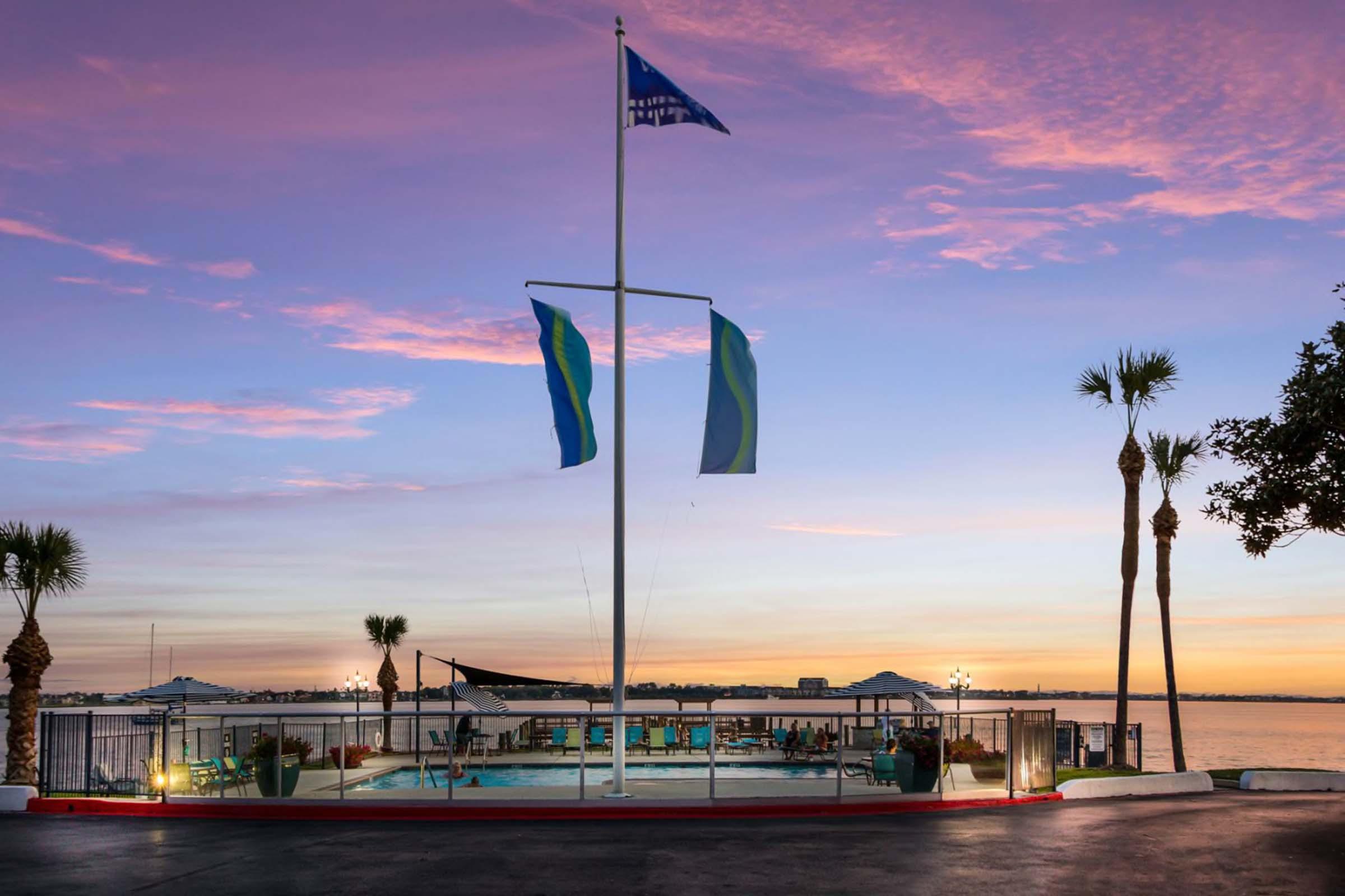 View of a waterfront facility at sunset, featuring a swimming pool surrounded by palm trees. Flags are flying atop a tall pole, and the sky is painted in shades of pink and blue. The calm water reflects the evening light, creating a serene atmosphere.