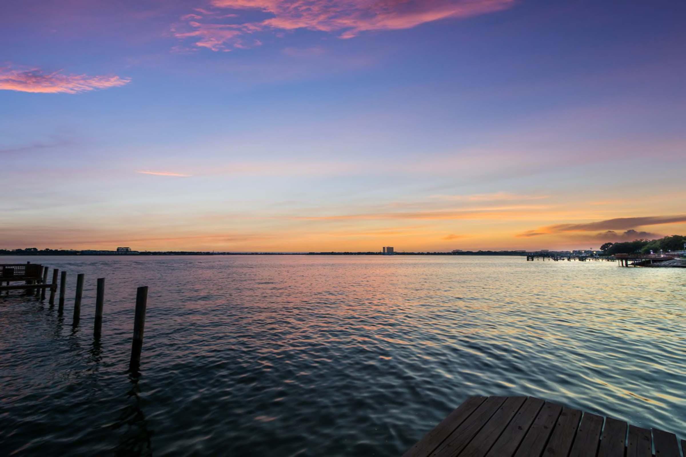 A serene waterfront scene at dusk, featuring calm waters reflecting pastel hues of purple, pink, and orange in the sky. Silhouettes of wooden posts jut out from the shore, and distant buildings are visible along the horizon, creating a peaceful, picturesque atmosphere.
