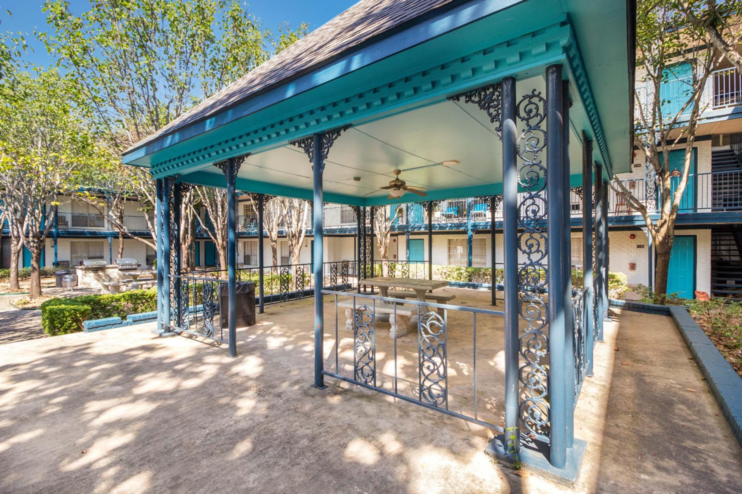 A scenic gazebo with ornamental ironwork and a ceiling fan, surrounded by trees. It is situated in an outdoor area next to a building with multiple units. Sunlight filters through the leaves, creating a bright and inviting atmosphere for relaxation or gatherings.