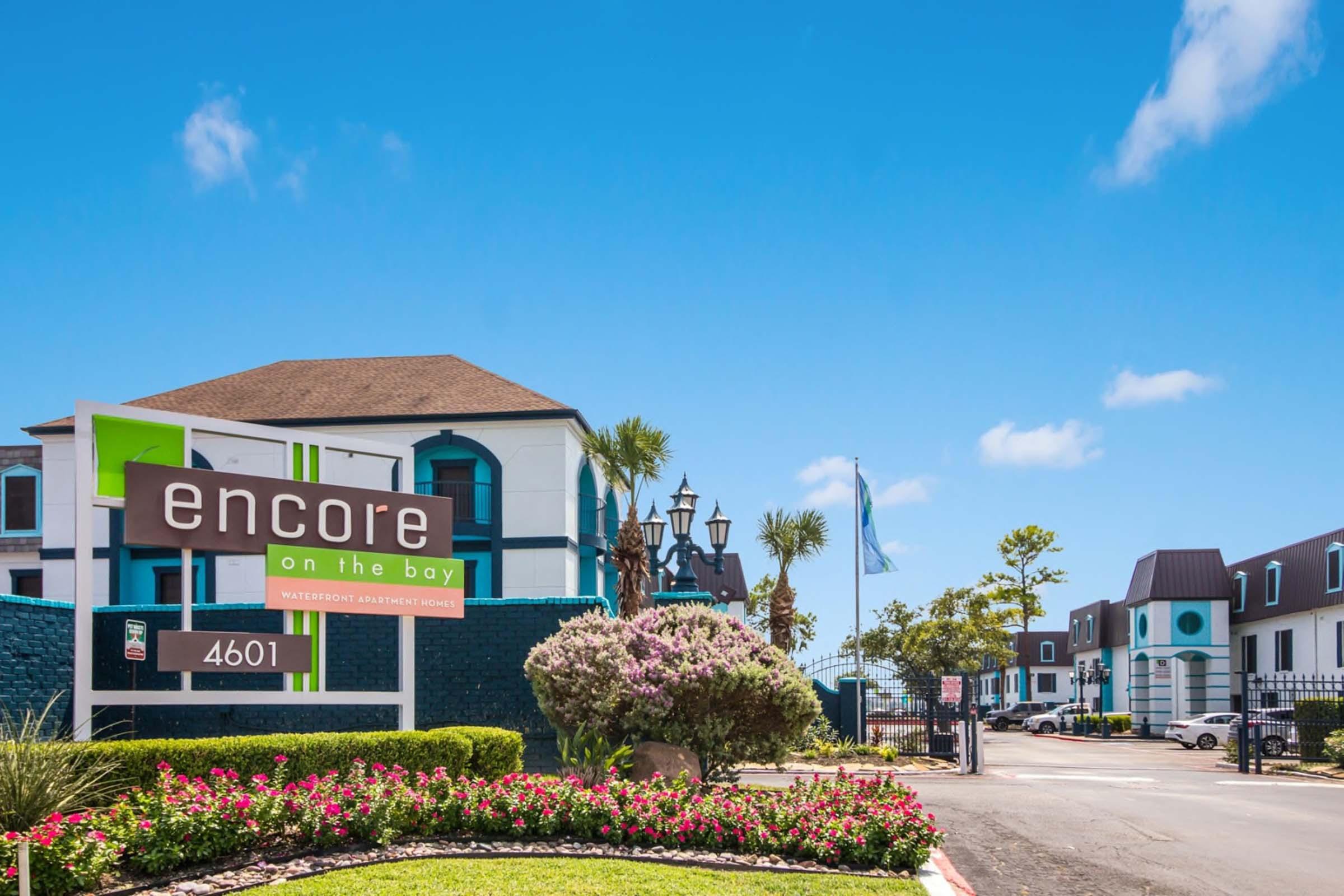 A colorful sign reading "encore on the bay" at the entrance of a waterfront apartment complex. The building features a mix of turquoise and white colors with palm trees and landscaping. Clear blue skies complete the vibrant atmosphere of the location.