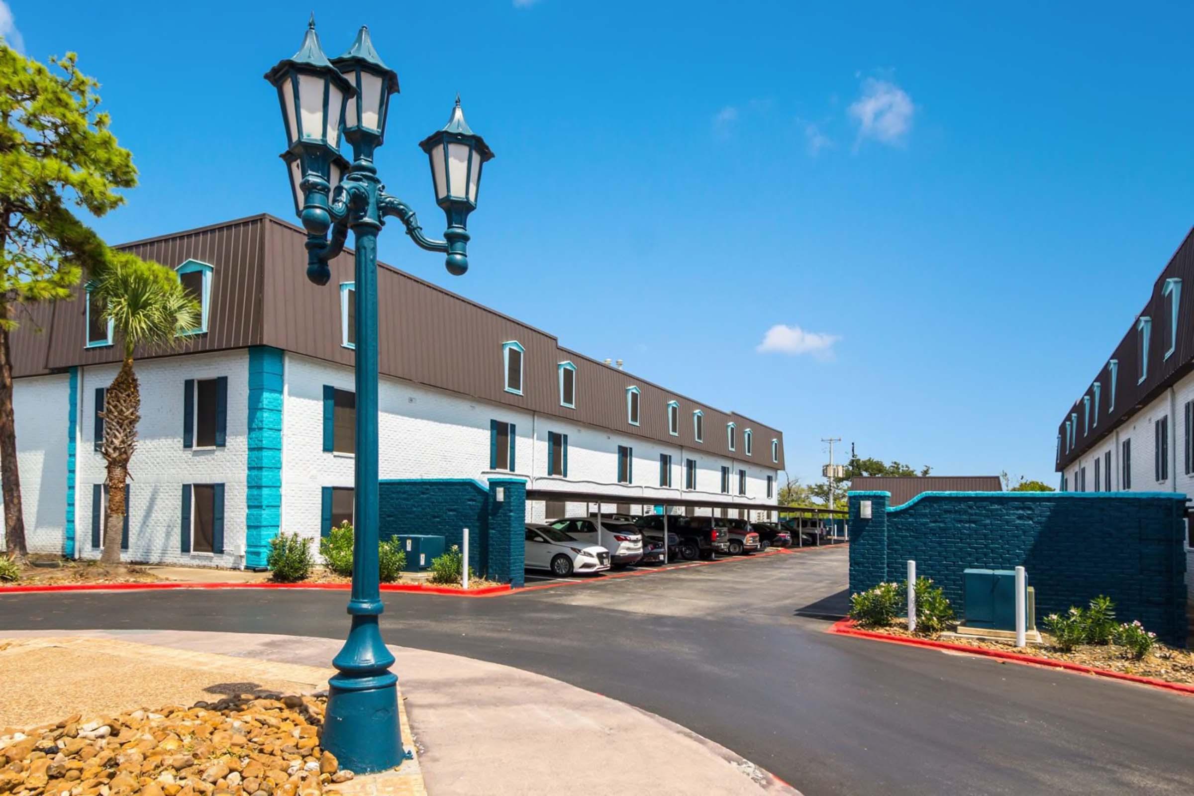 Exterior view of a hotel featuring a turquoise and white color scheme. There is a decorative lamp post in the foreground and parked cars under a covered area. Lush green landscaping surrounds the building, and the sky is clear with a few clouds. The hotel has two levels and shows a welcoming atmosphere.