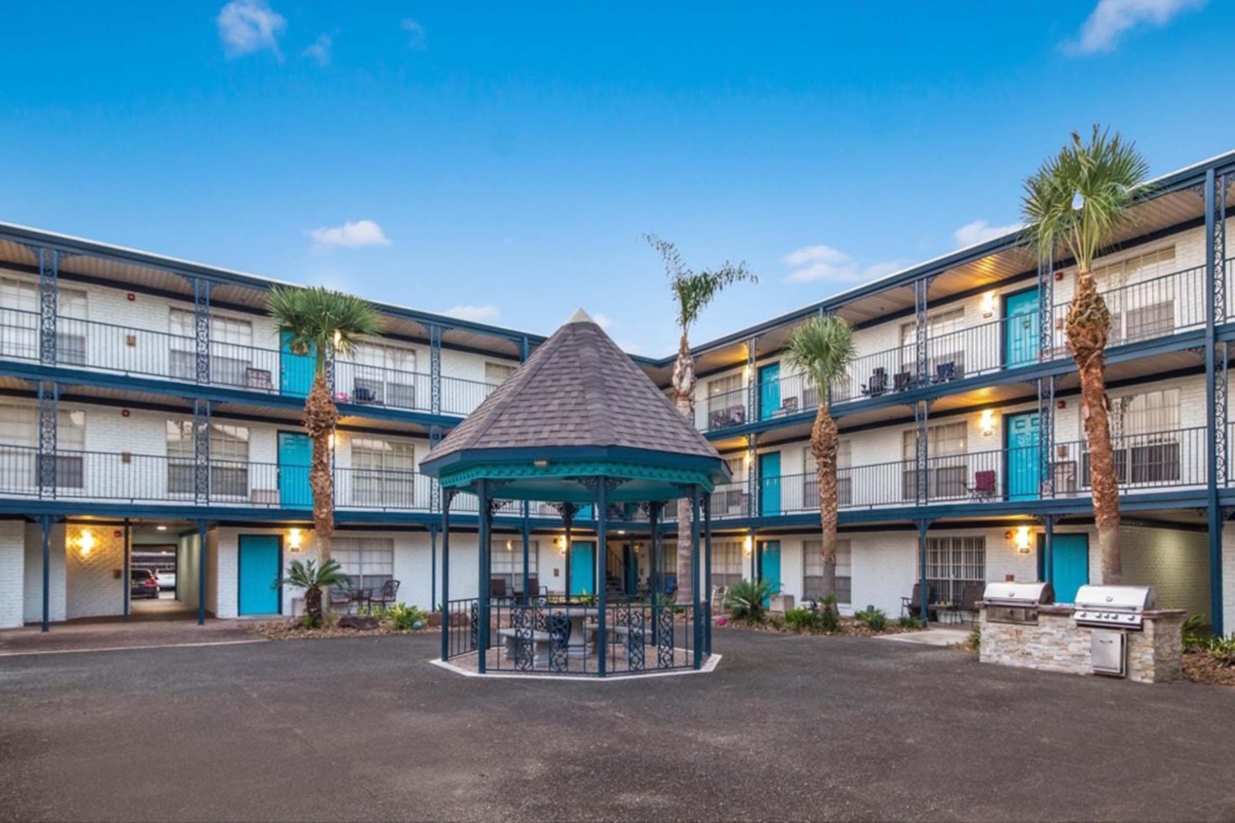 A courtyard view of a hotel with a gazebo at the center. Surrounding the gazebo are palm trees and grill stations. The building features multiple floors with blue doors and balconies, set against a bright blue sky.