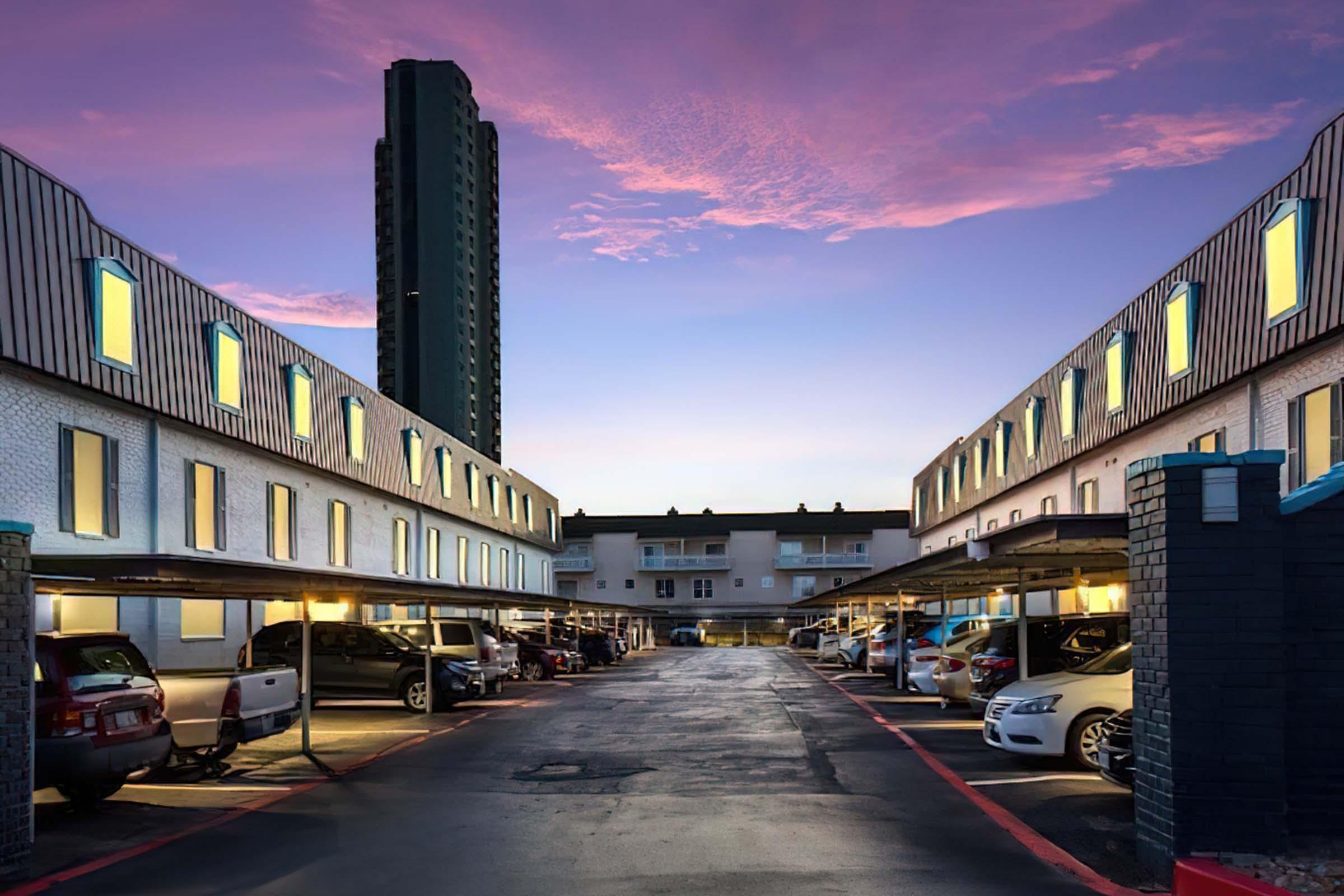 View of a modern motel or apartment complex at twilight, featuring two parallel buildings with illuminated windows. The foreground shows covered parking spaces with several cars, while a tall building stands in the background under a colorful sky with pink and purple hues.