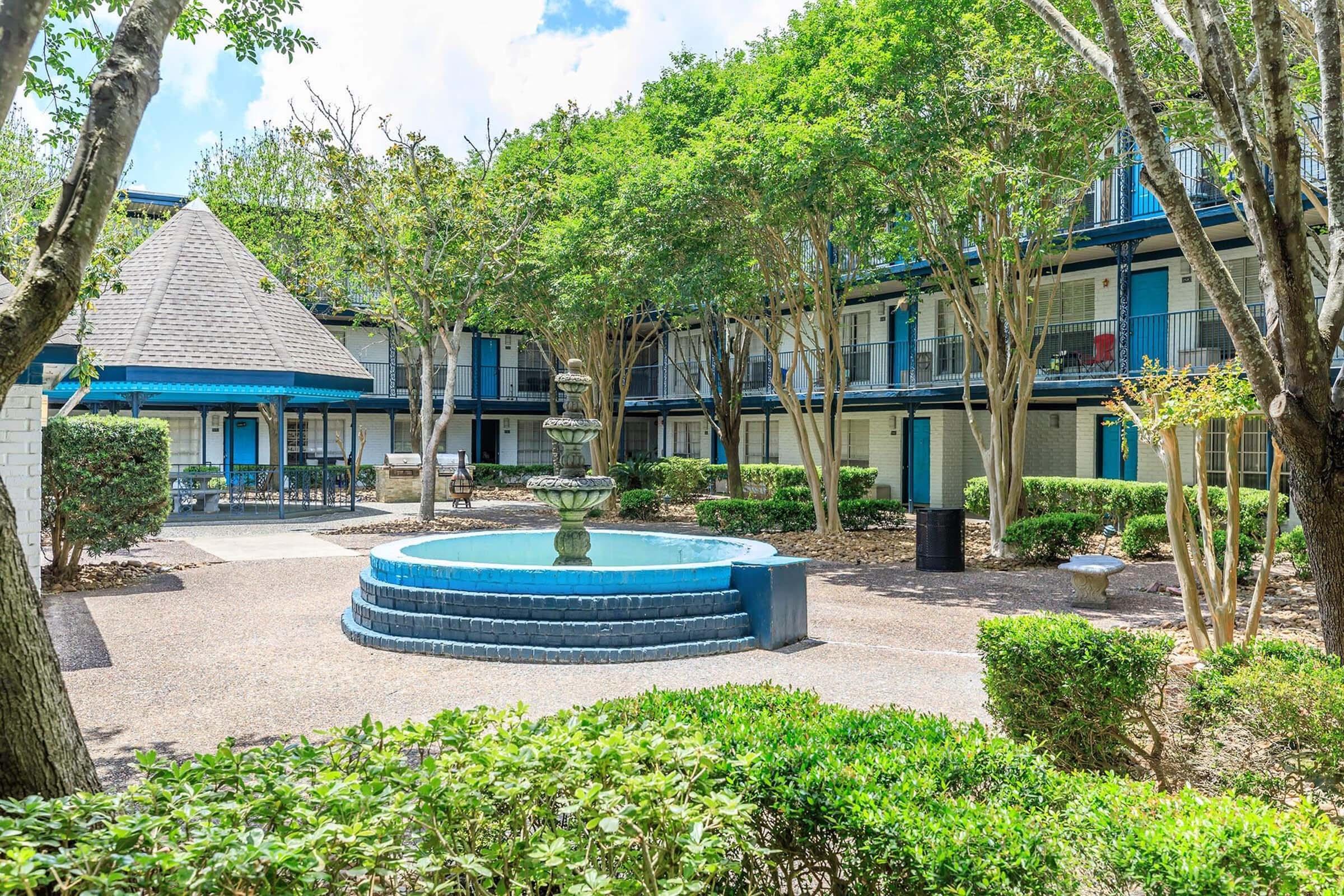 A well-maintained courtyard featuring a blue fountain surrounded by lush greenery and trees. The area includes paths and seating, with low shrubs and a mix of light and shaded spaces. Two-story buildings can be seen in the background, creating a tranquil atmosphere.
