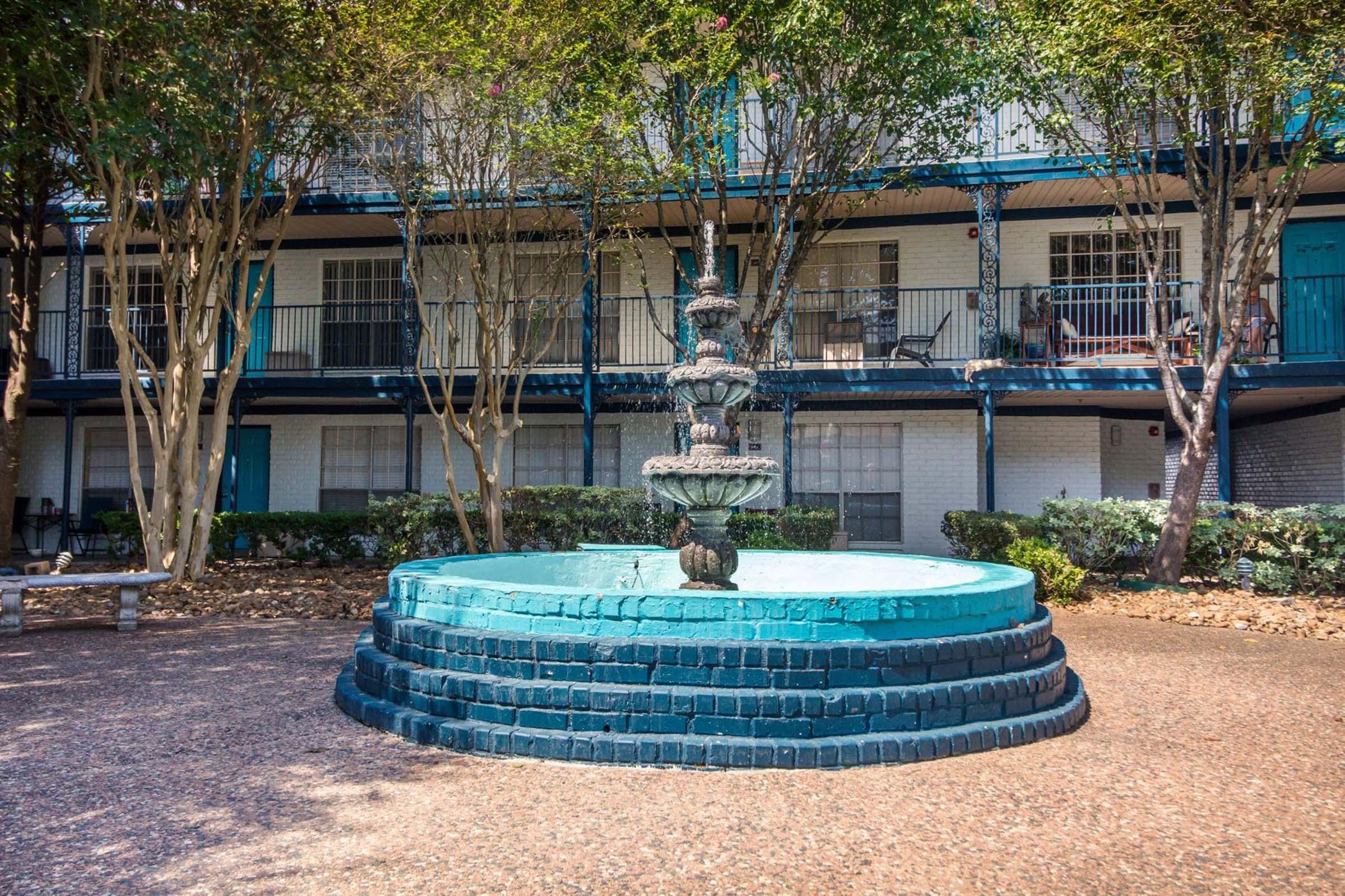 A decorative fountain in a courtyard surrounded by low-rise apartment buildings. The fountain features multiple tiers of water spouts, with blue paint highlighting its structure. Lush greenery and seating areas are present, creating a serene outdoor space.