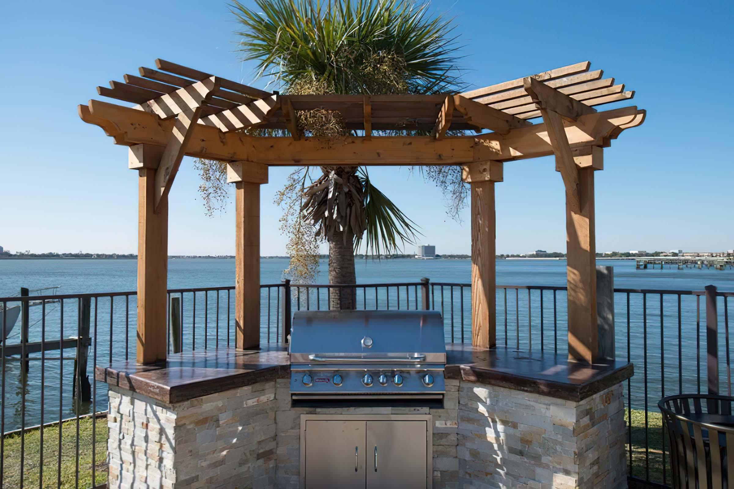 An outdoor grilling station under a wooden pergola, featuring a stainless steel barbecue grill. In the background, there is a scenic view of a body of water and a clear blue sky, with a palm tree nearby. The area is enclosed by a black railing.