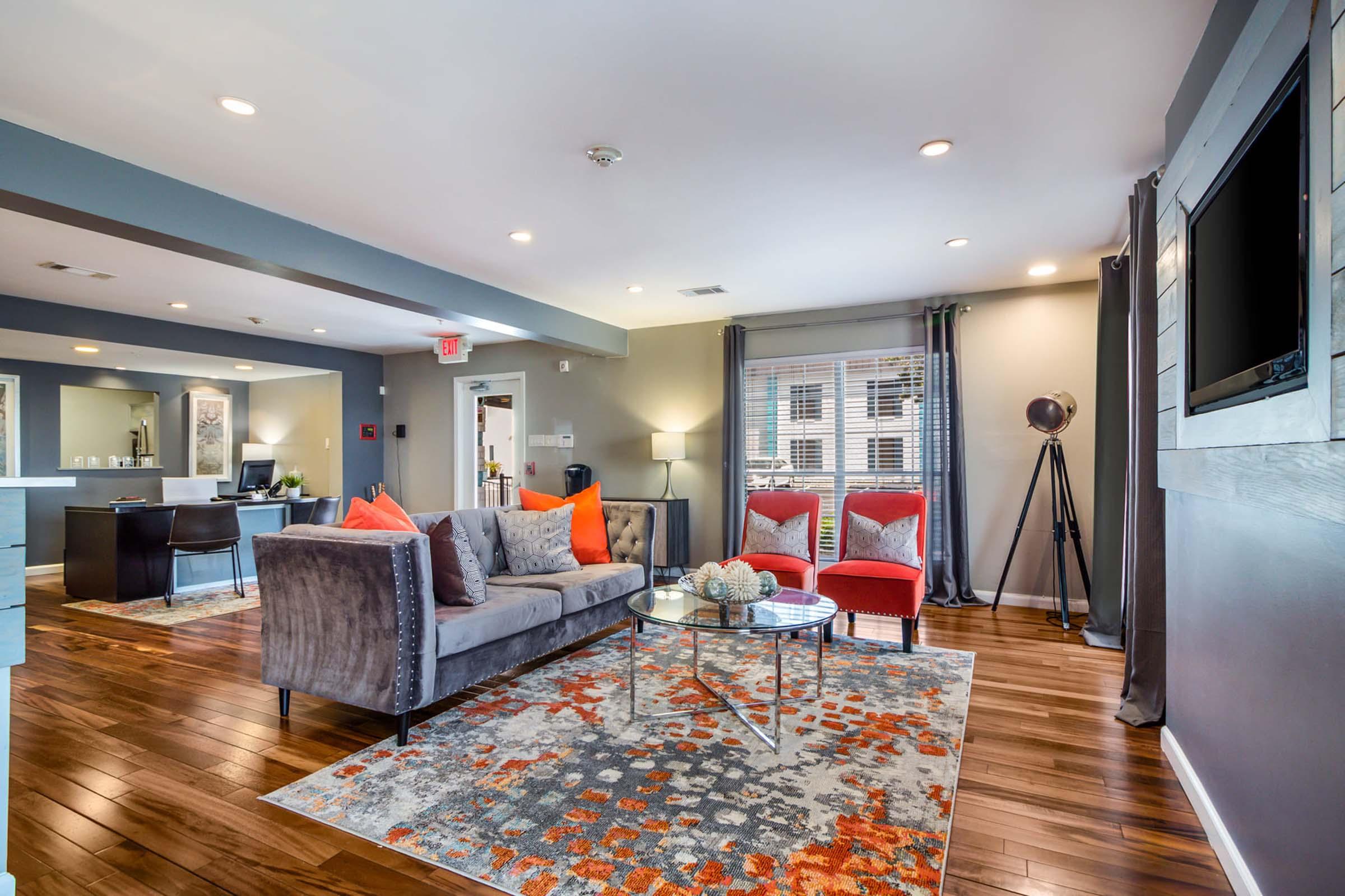 Modern living room with a gray sofa, two red accent chairs, and a glass coffee table on a vibrant rug. The walls are painted a soft gray, and there are large windows allowing natural light. Decorative items are placed on the table, and a sleek floor lamp stands in one corner.