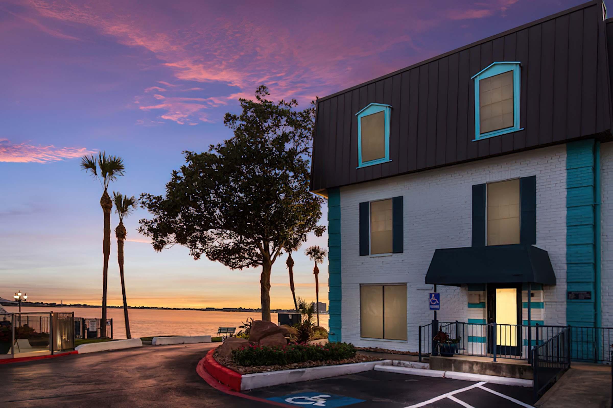 A coastal hotel at dusk with a vibrant pink and purple sky. The building features a mix of white and teal walls, palm trees nearby, and a pathway leading to the water's edge. The scene conveys a tranquil, scenic ambiance perfect for relaxation by the water.
