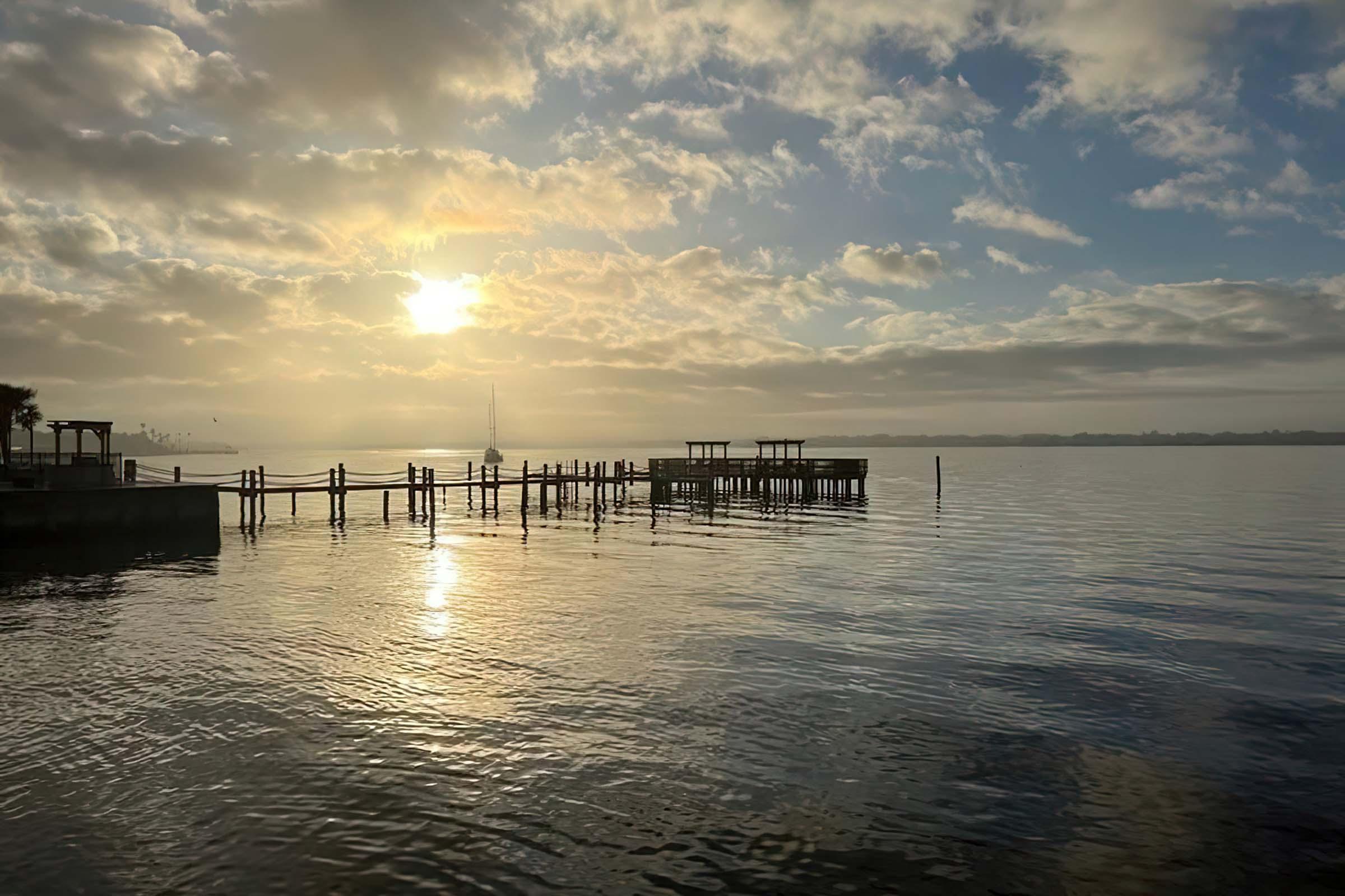 A serene view of a calm body of water at sunrise, reflecting golden hues. A wooden dock extends into the water, surrounded by a soft mist. Clouds drift across the sky, adding depth to the scene. The atmosphere feels tranquil and peaceful, suggesting an early morning by the waterfront.