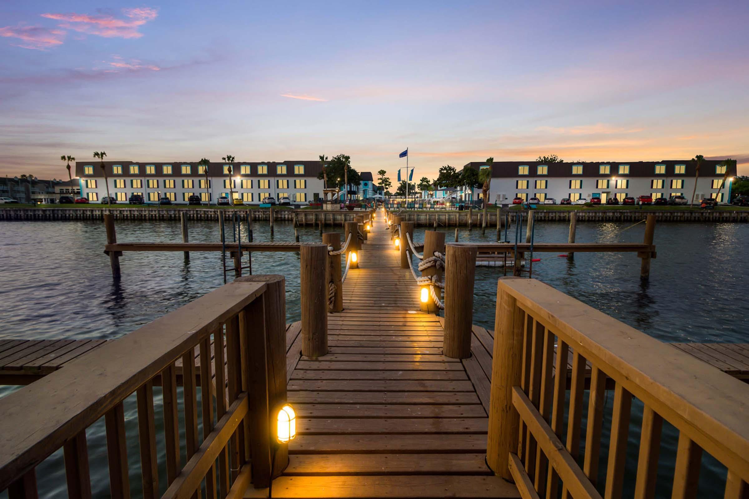 A wooden pier extends into calm water, lined with lights on either side. In the background, buildings with multiple windows reflect the sunset colors in the sky. The serene setting creates a peaceful atmosphere, ideal for a tranquil evening.