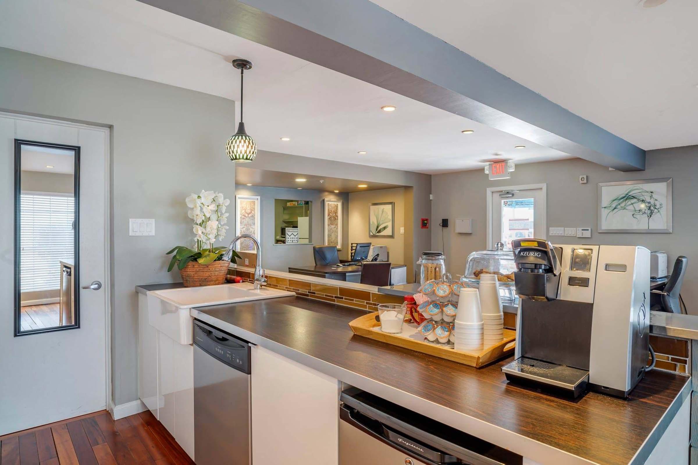 Modern kitchen with a focus on a coffee station. Features a countertop with a sleek coffee machine, glass jars with snacks, and a vase with white flowers. Soft lighting from overhead fixtures creates a warm atmosphere. The area includes a mirror and a doorway leading to another space.