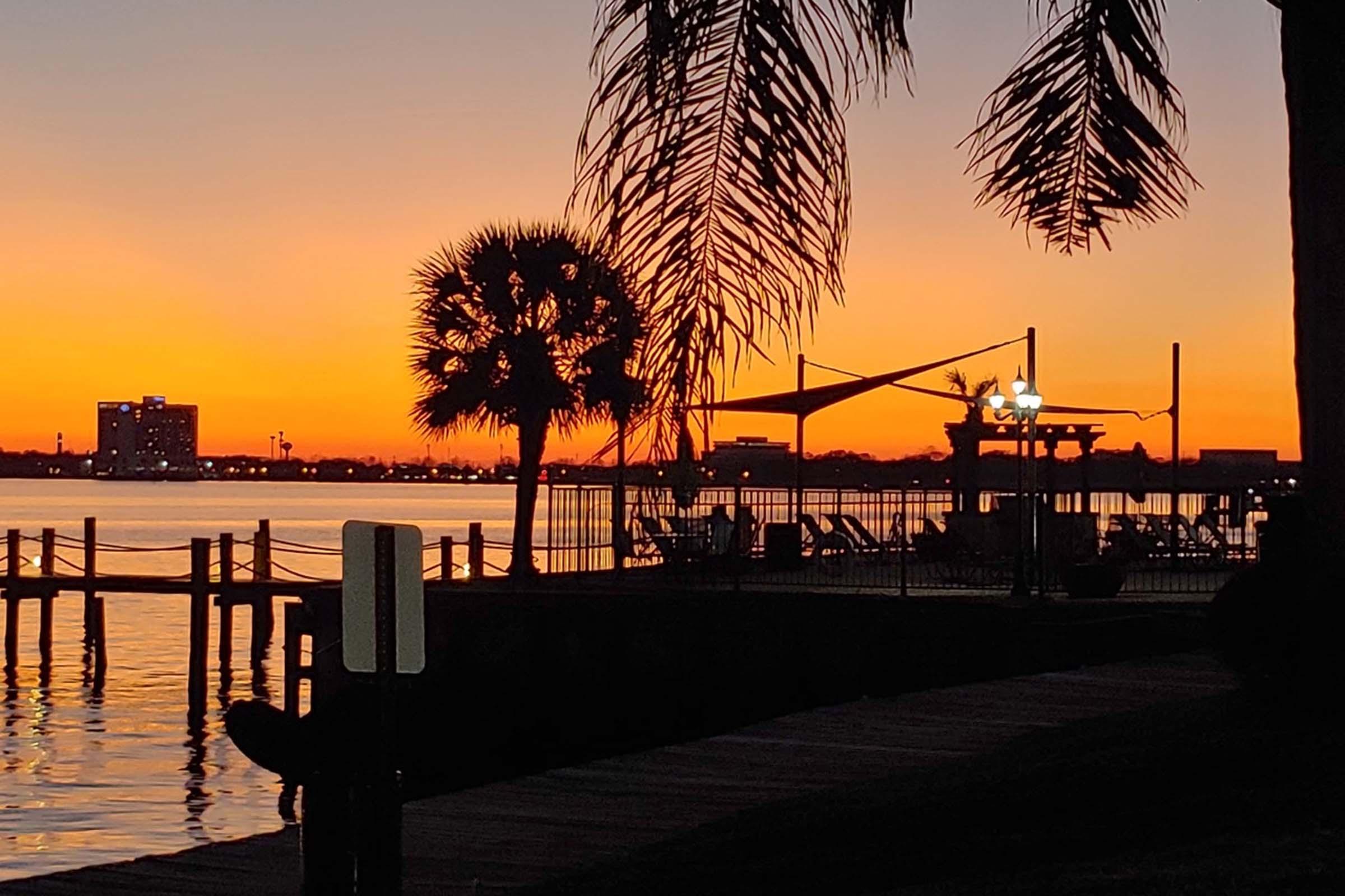 A serene sunset over a calm body of water, with silhouettes of palm trees and a pier in the foreground. The sky is painted in vibrant shades of orange and purple, while lights near a dock illuminate the scene, creating a tranquil ambiance.