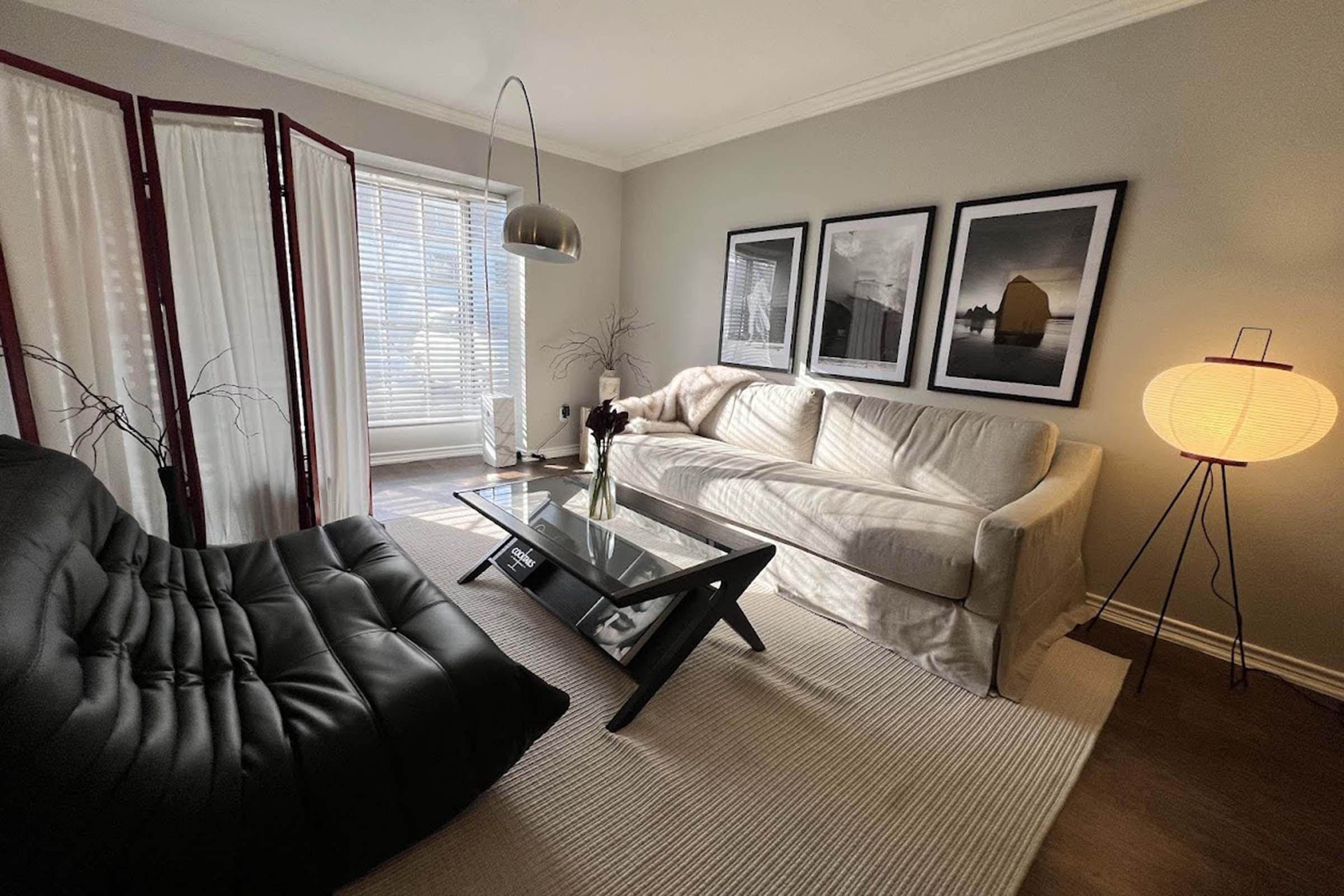A cozy living room featuring a light-colored sofa and a black, modern armchair. There is a glass coffee table in the center, surrounded by soft, neutral decor. Three framed black-and-white pictures hang on the wall, and a floor lamp with a round shade adds warmth to the space, complementing the natural light coming through the window.
